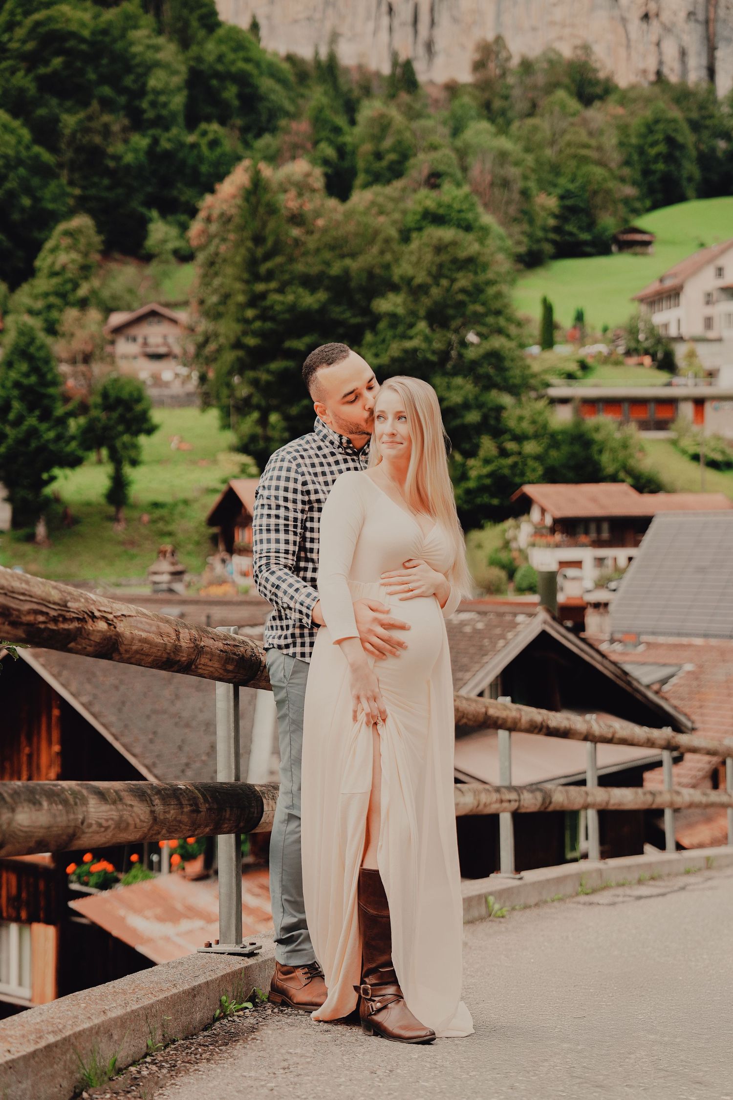 Couple posing together on bridge in Swiss alpine village, man kissing pregnant partners head tenderly.