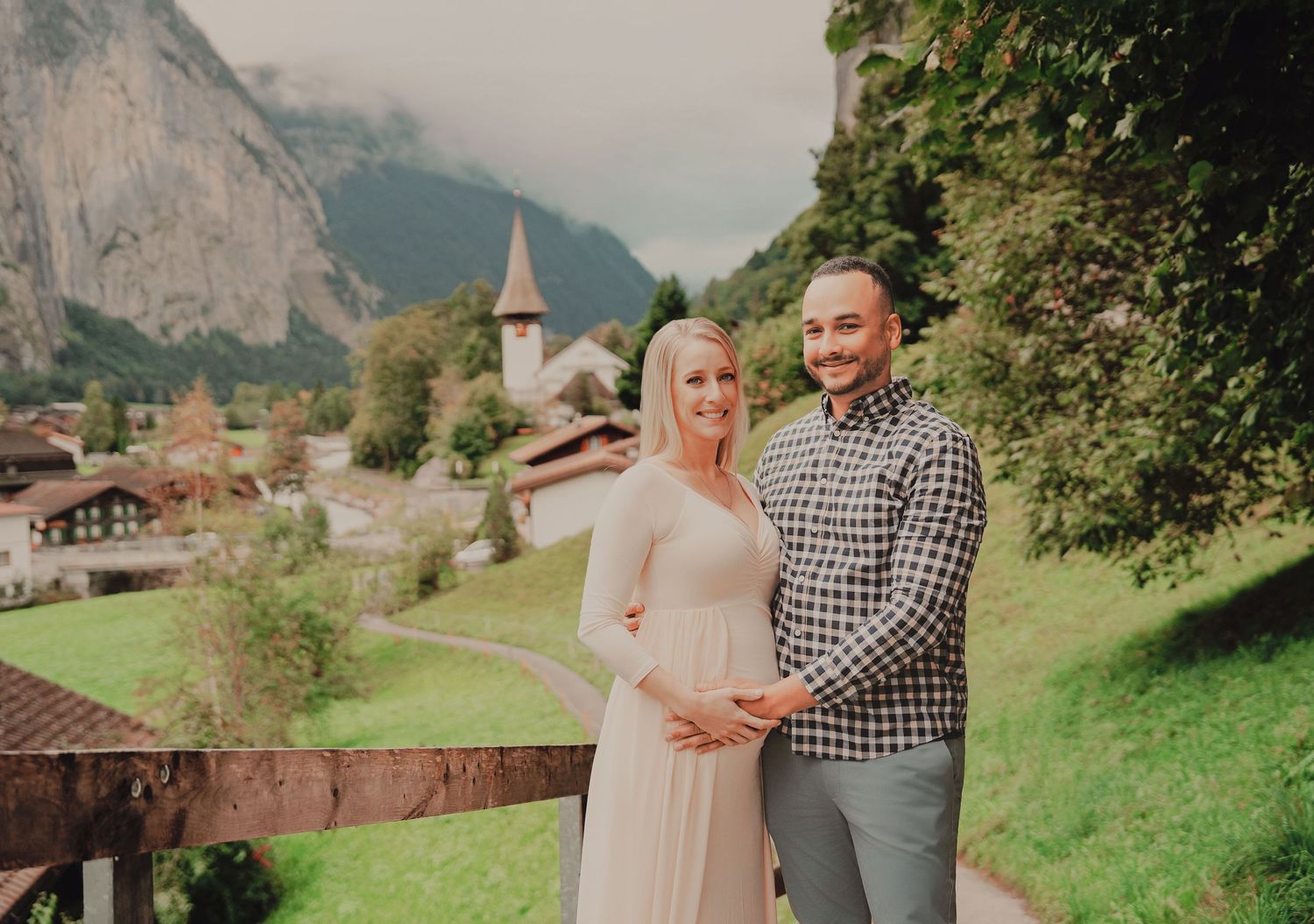 Smiling pregnant couple poses together on a balcony overlooking a lush Swiss alpine valley village.