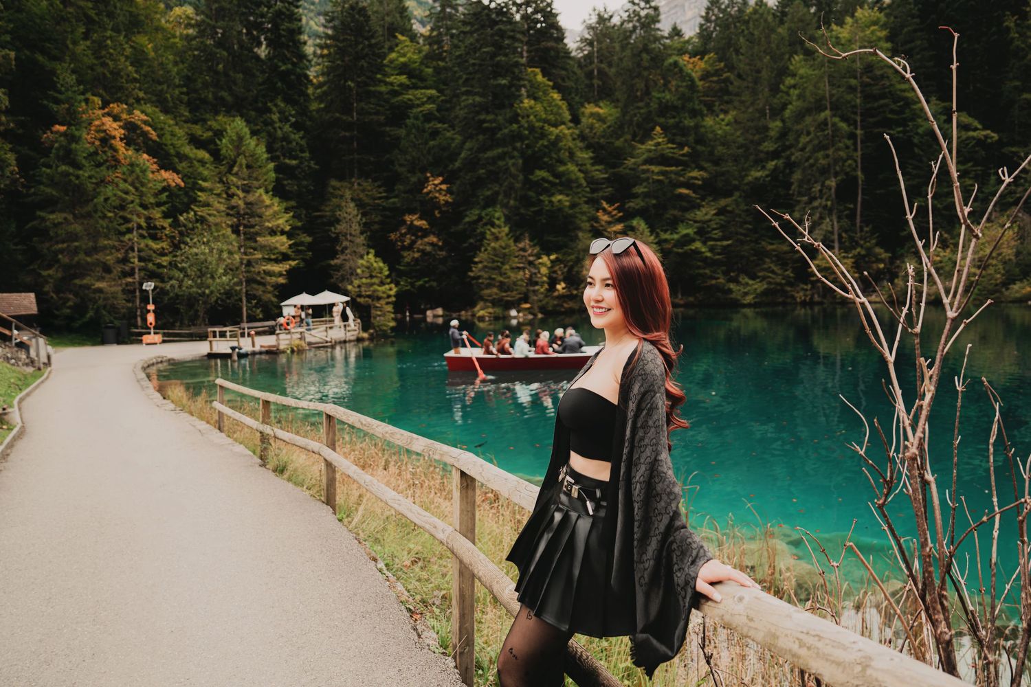 Smiling woman with long red hair in black outfit posing by stunning turquoise Blausee lake in Switzerland.