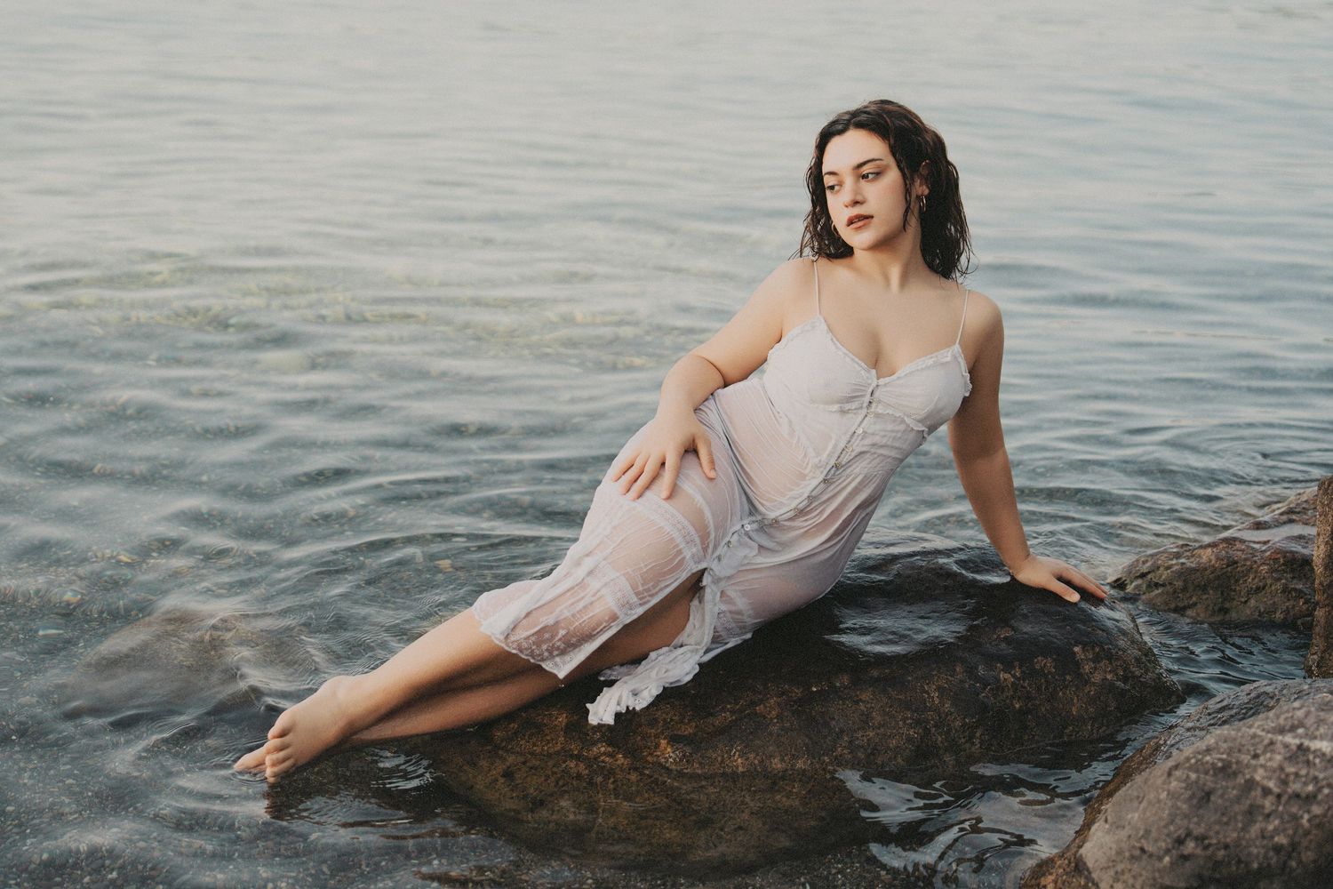 Woman in sheer white dress sitting gracefully on rocks by calm sea.
