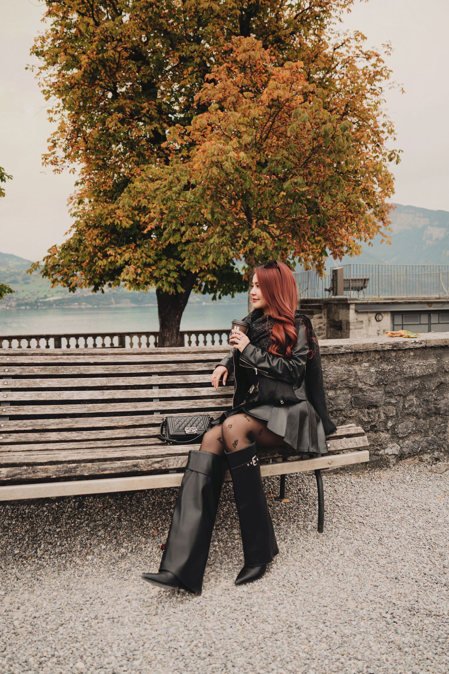 Stylish redhead woman in black leather jacket sitting on bench under golden autumn tree.