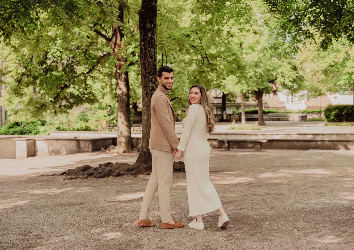 Couple holding hands and looking back at camera in a lush green park, woman in white dress, man in tan blazer.
