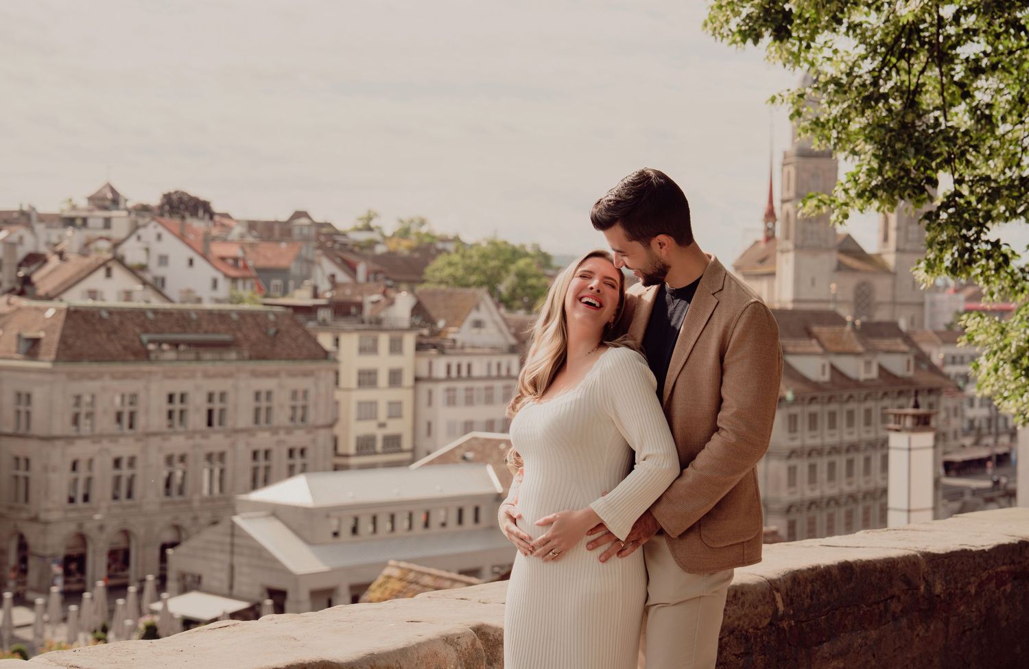 Happy pregnant couple laughs together on stone terrace overlooking Zurich old town rooftops and church spires.