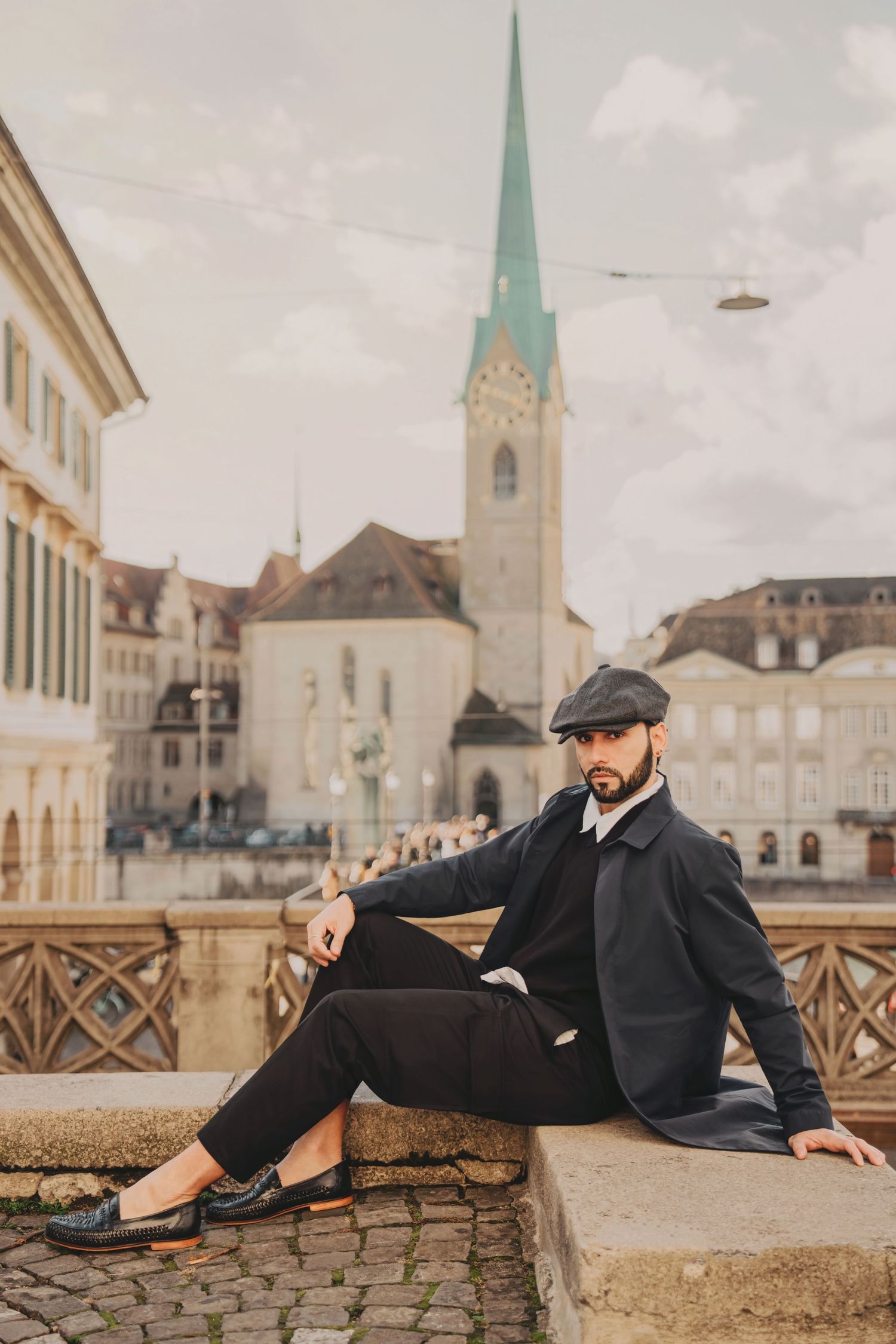 Well-dressed bearded man in dark coat and flat cap sits relaxed on stone ledge in historic Zurich city center.
