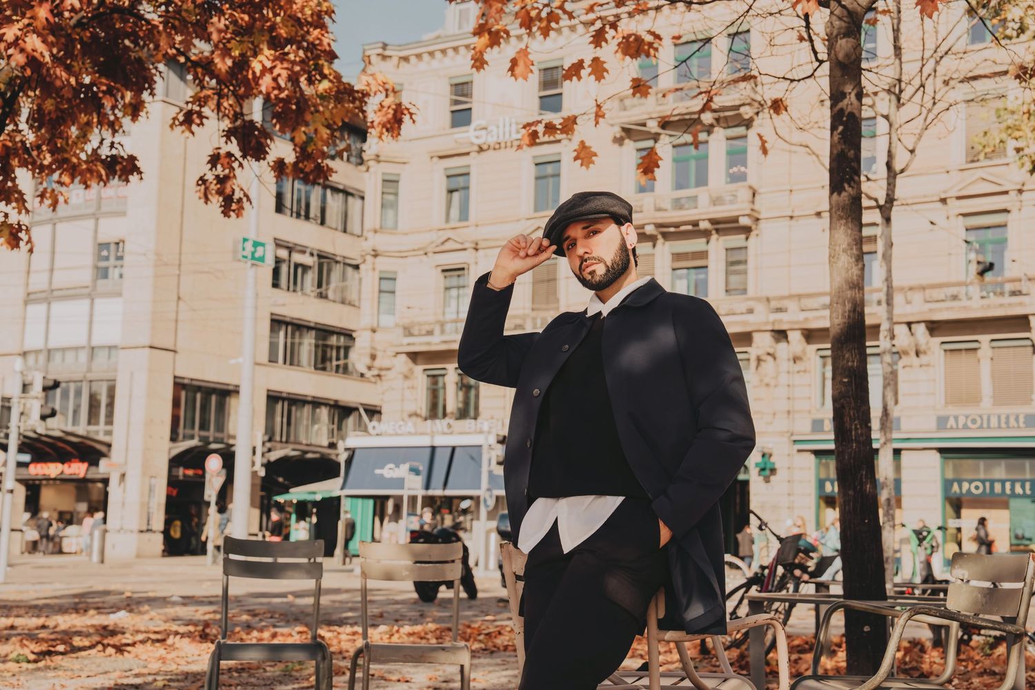 Bearded man in elegant black coat and newsboy cap standing on vibrant autumn city street.