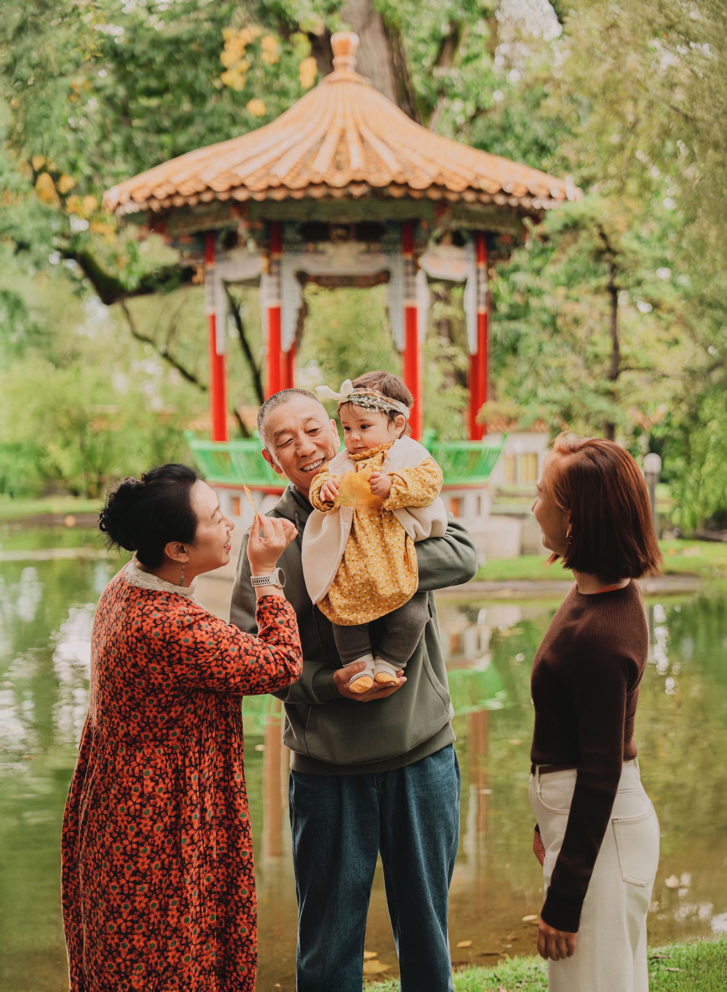 Three generations of Asian family laughing and playing with baby near Chinese pavilion and reflective garden pond.