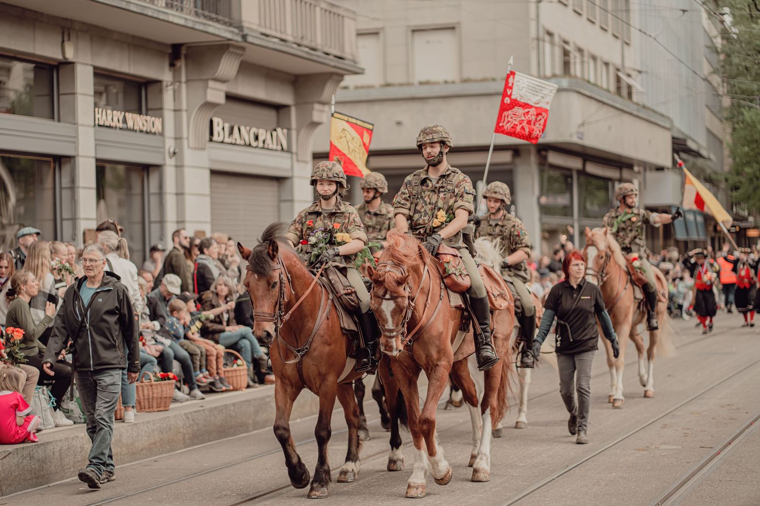 Military riders on horseback carrying colorful flags parade through a crowded city street.
