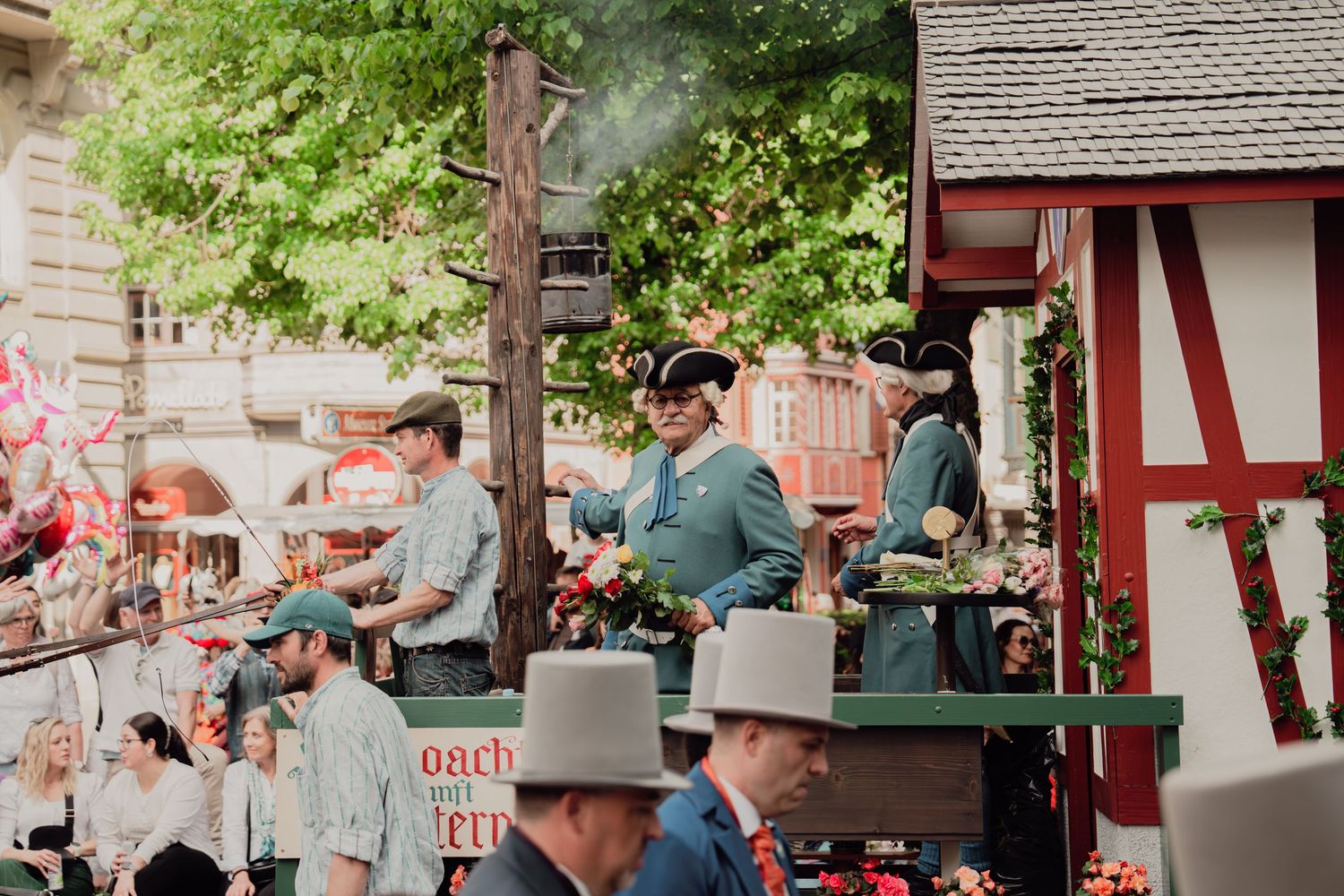 Historical reenactors in period military uniforms ride a decorated parade float past a traditional half-timbered building in summer.