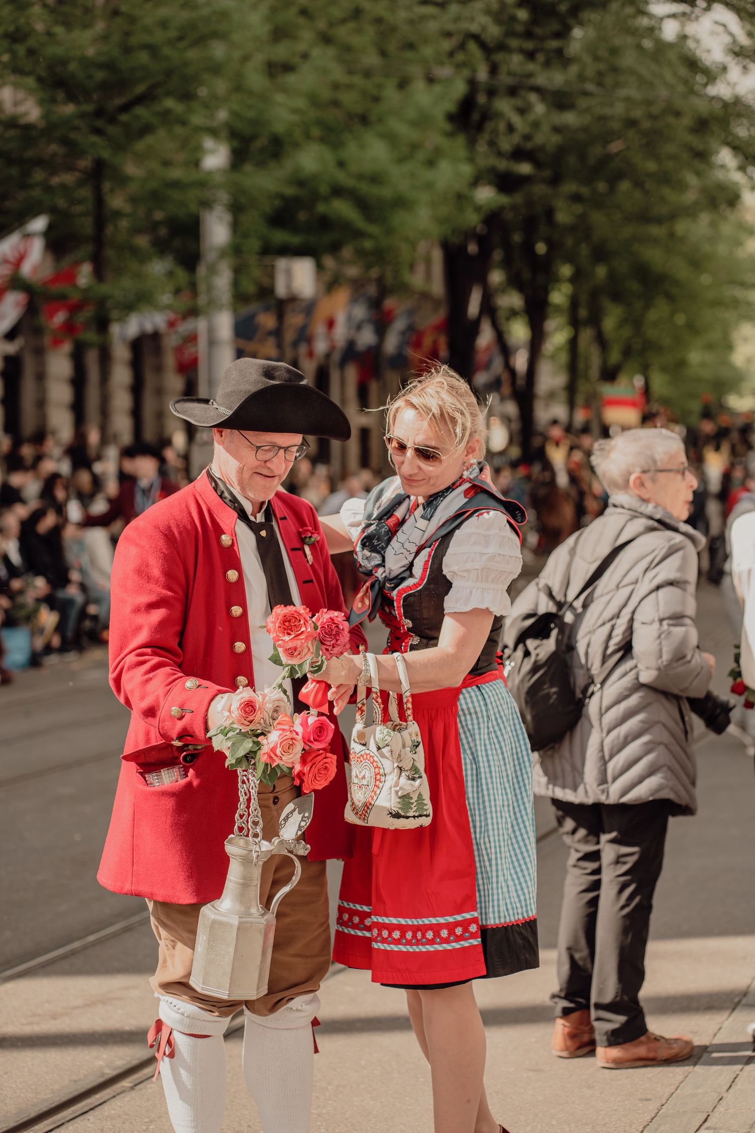Man in red 18th-century coat offers roses to woman in traditional dirndl during a festive street parade.