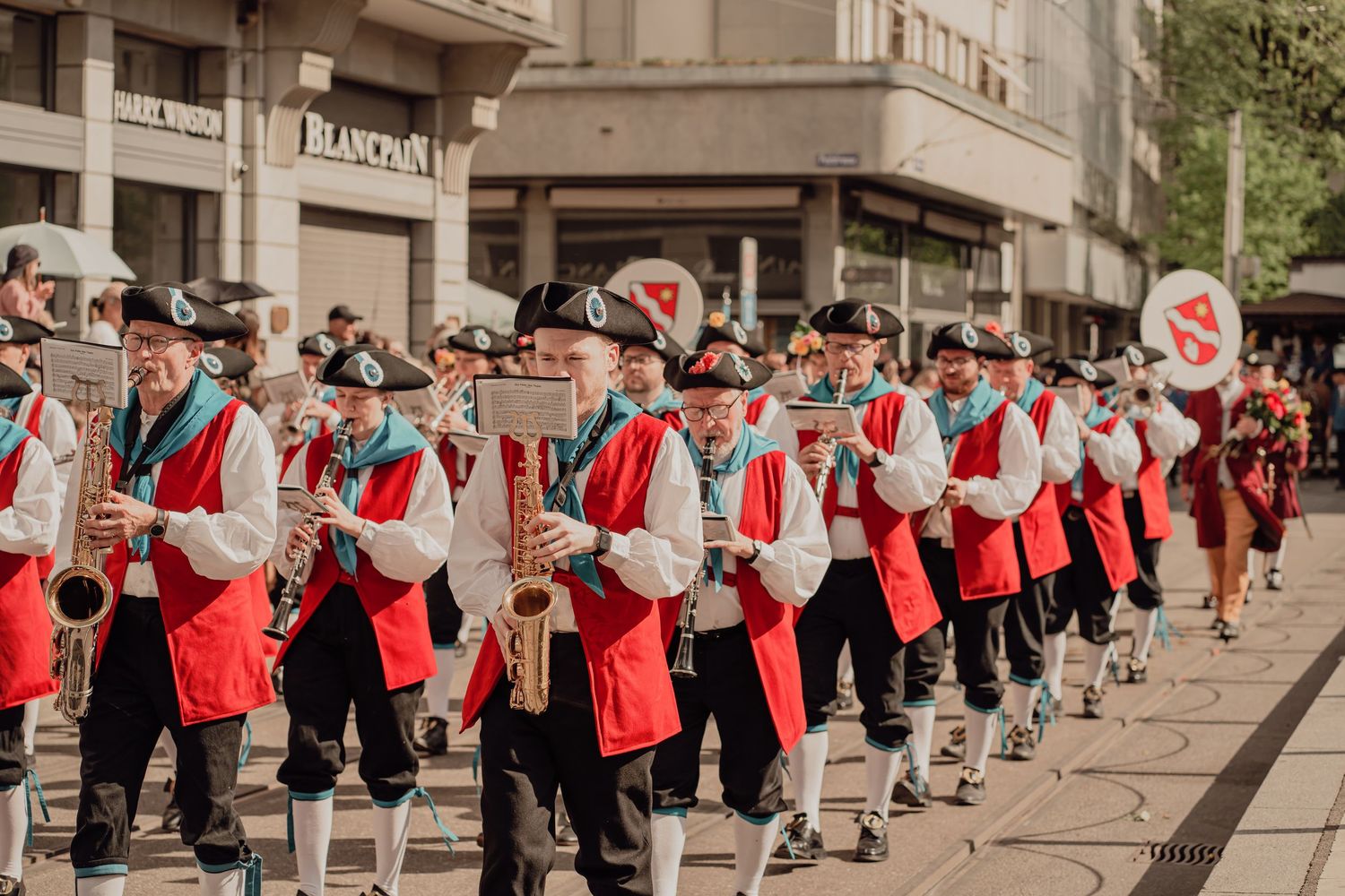 Traditional marching band in red and black uniforms playing saxophones during a street parade.