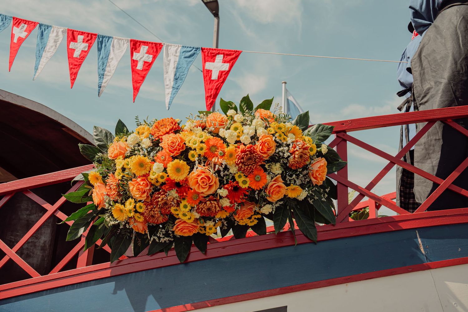 Vibrant orange and yellow rose floral arrangement on a red railing decorated with Swiss flags and colorful bunting.