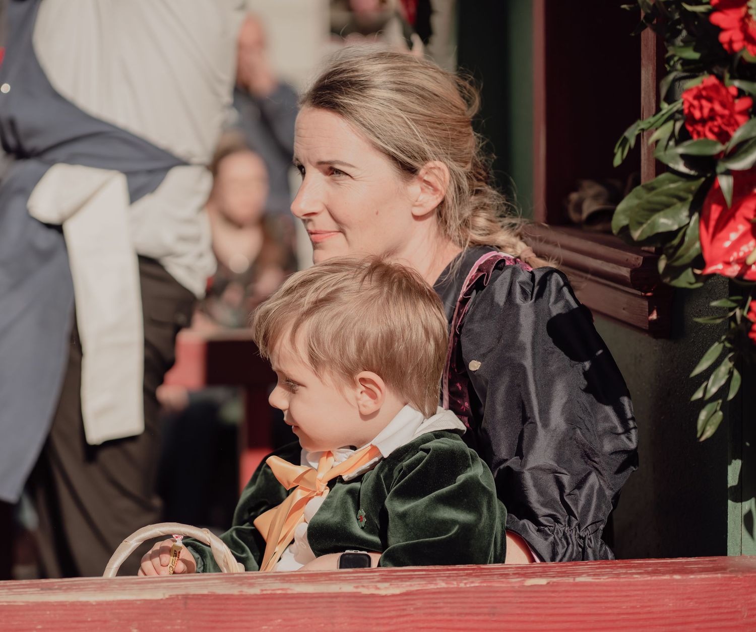 A blonde woman and young child in period costume sit together on a decorated red parade float.