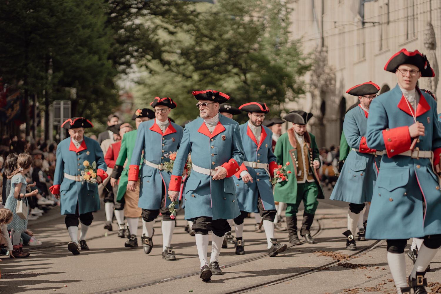 Men in matching blue and red 18th-century military uniforms march proudly in a street parade.