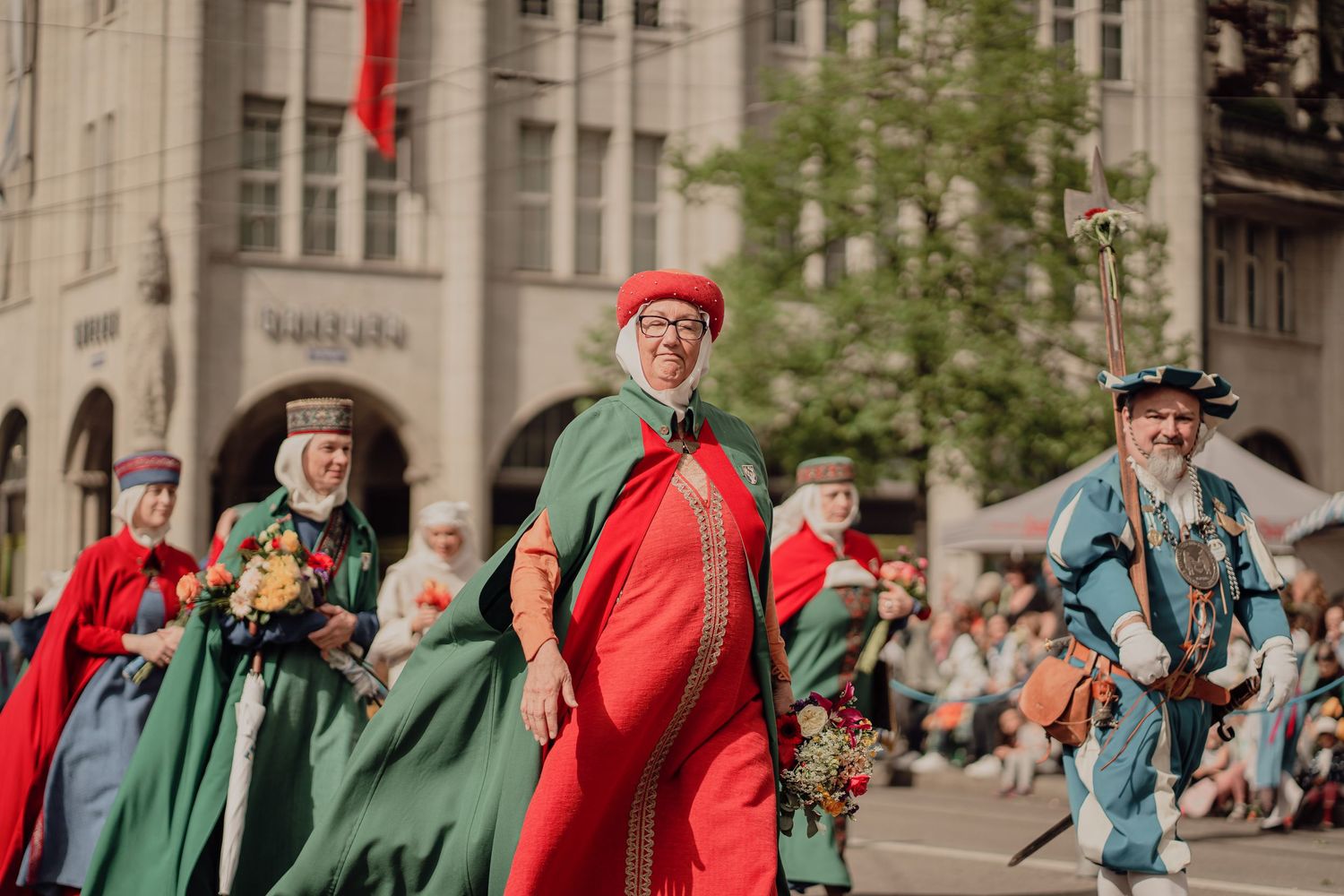 Participants in colorful red and green medieval costumes march through a city street during a festive parade.