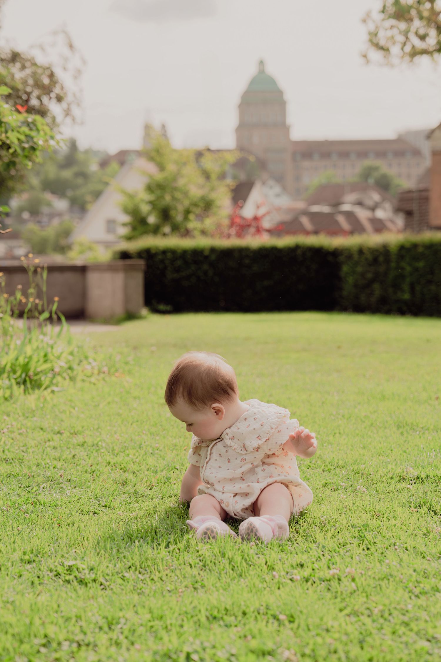 Baby in floral romper sitting on green grass in a garden with historic building backdrop.
