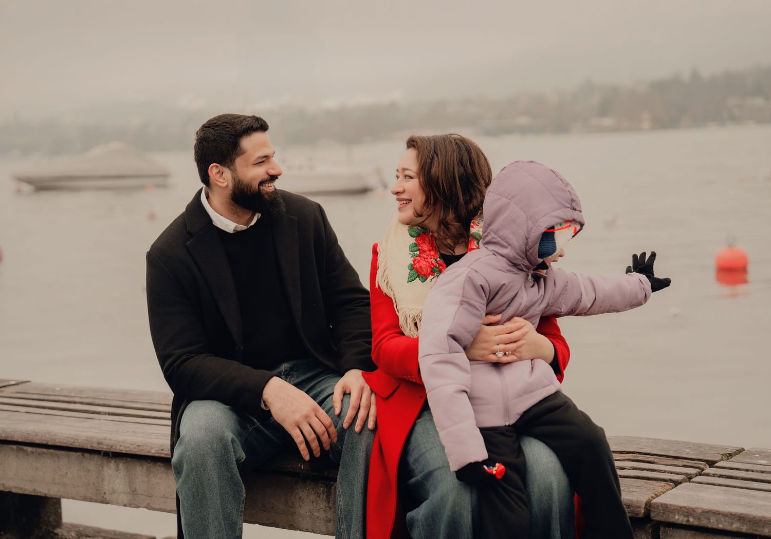 Happy family sits on a dock by a misty lake, parents laughing as toddler reaches out playfully.