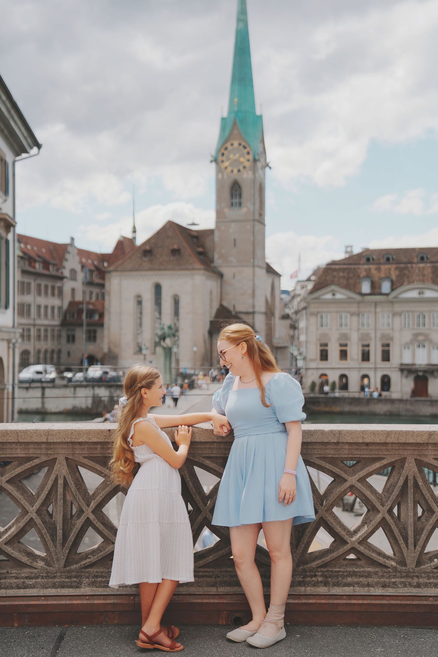 A mother and daughter in blue and white dresses stand on a bridge with Zurich old town church behind them.