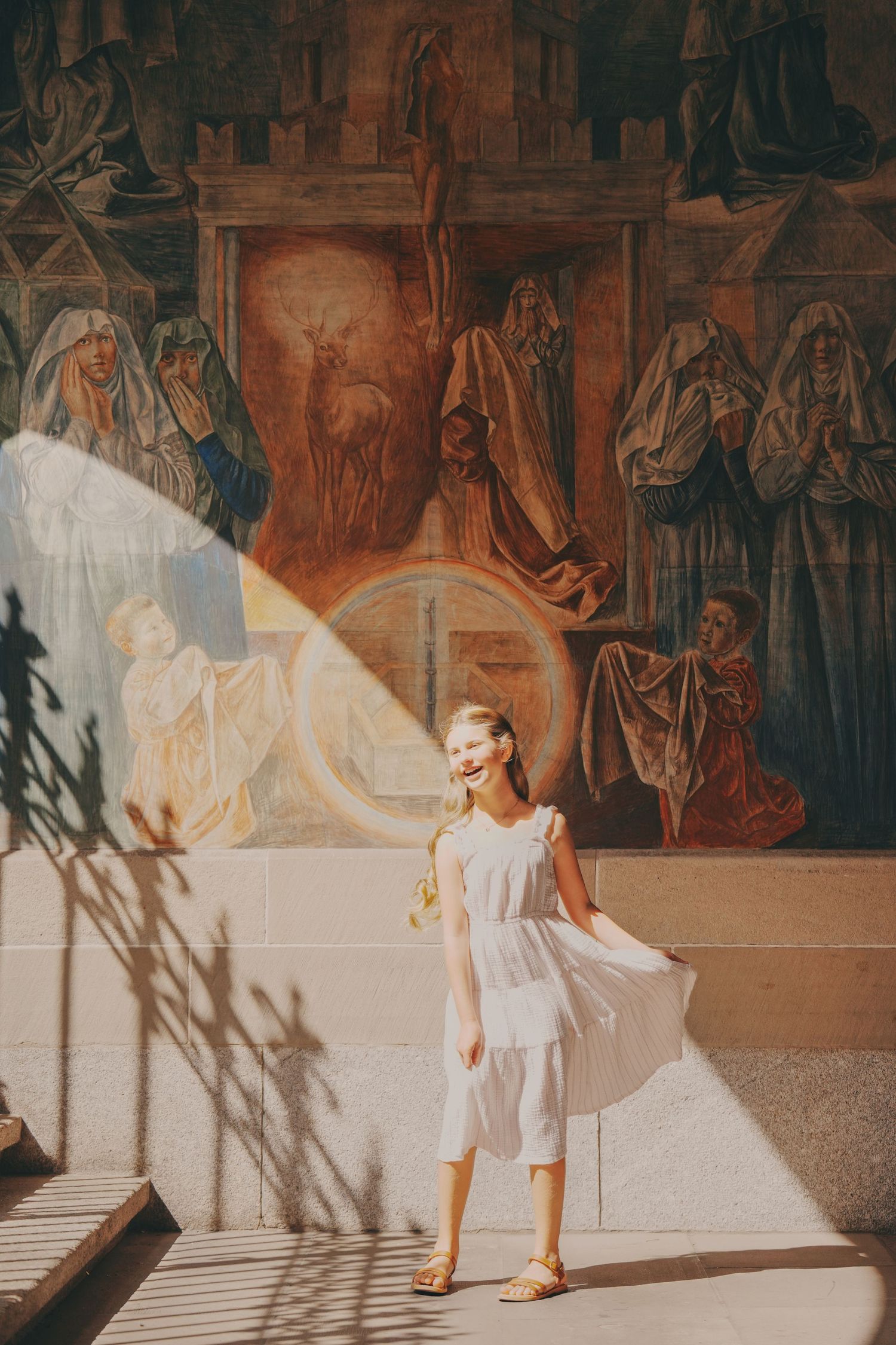 Happy girl in white dress standing before ornate religious mural in sunlit church entrance.