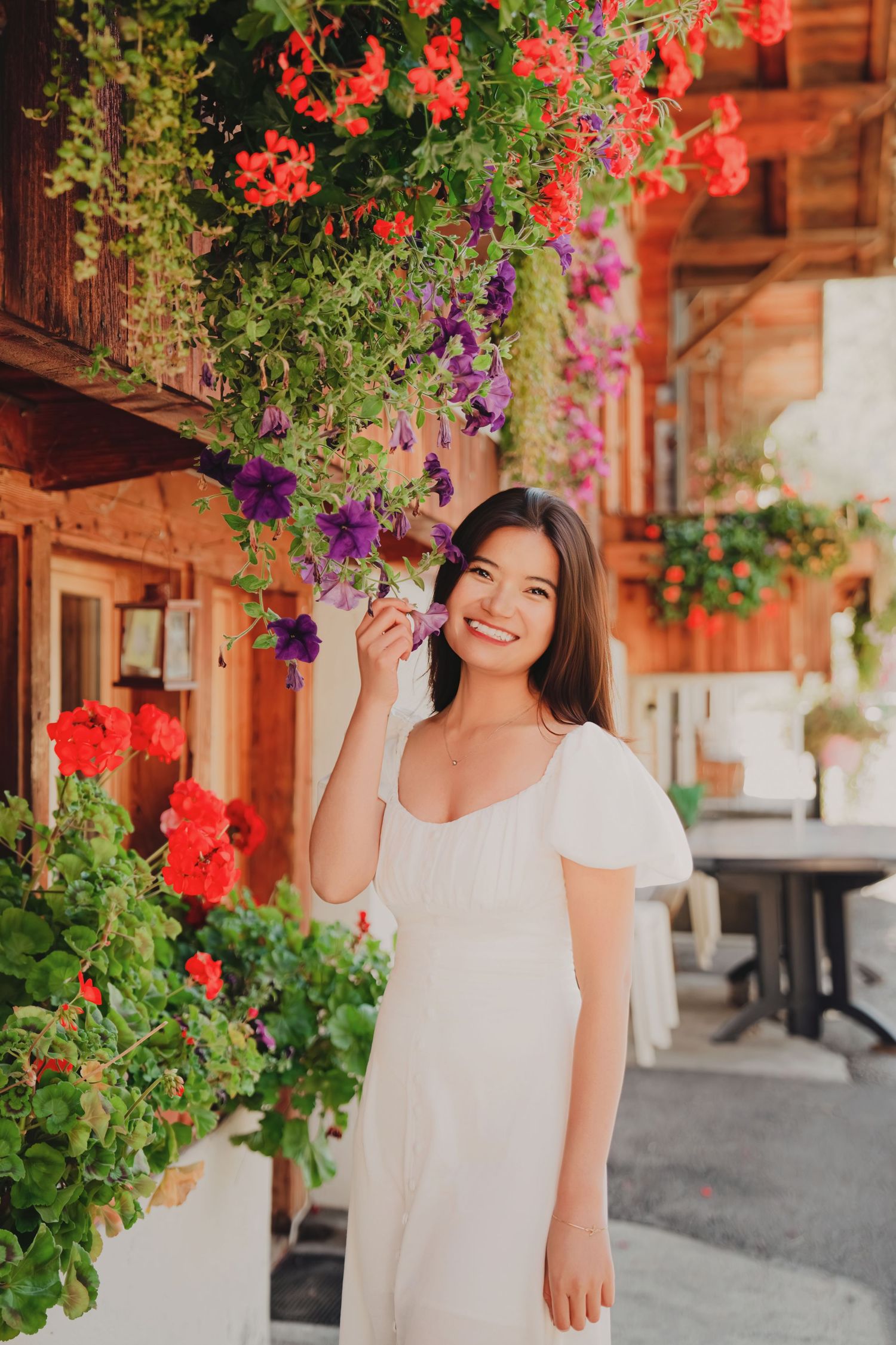 Beautiful woman in a white dress smiling while touching vibrant purple and red hanging flowers on a rustic wooden building.