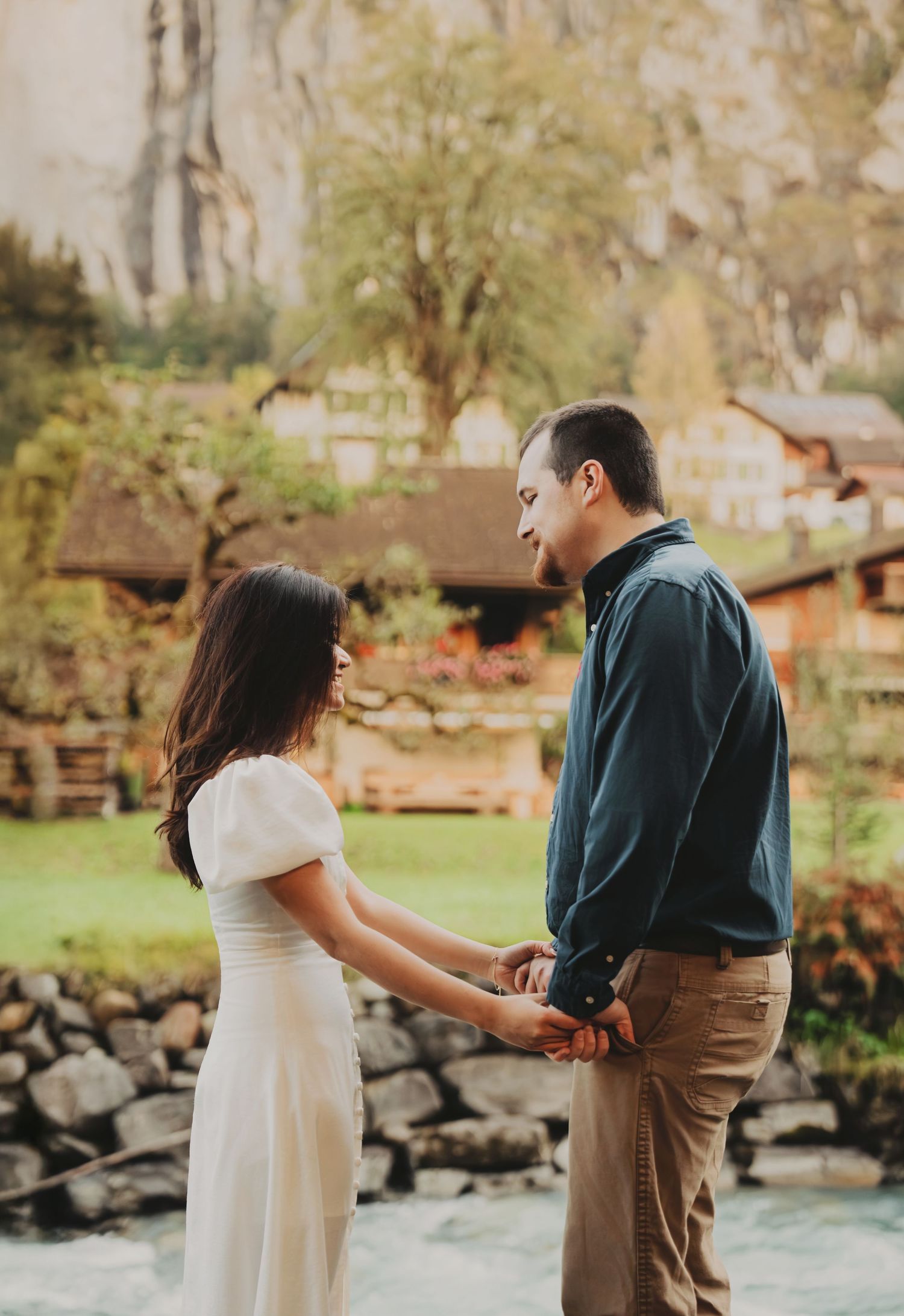 Couple holding hands by a river with Swiss alpine village and mountains in background.