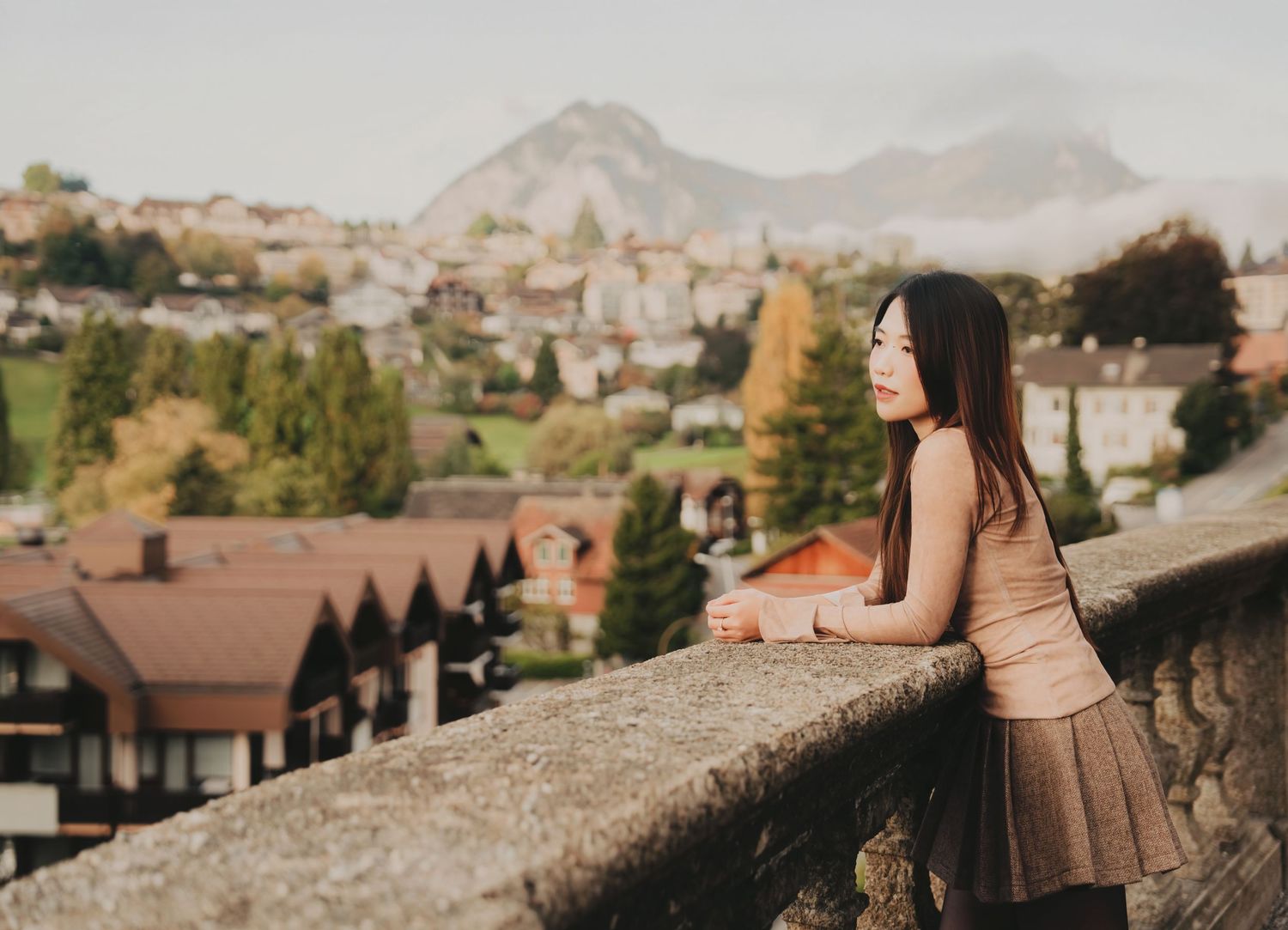 Woman in a beige dress leans on a stone wall enjoying panoramic views of a Swiss mountain village.