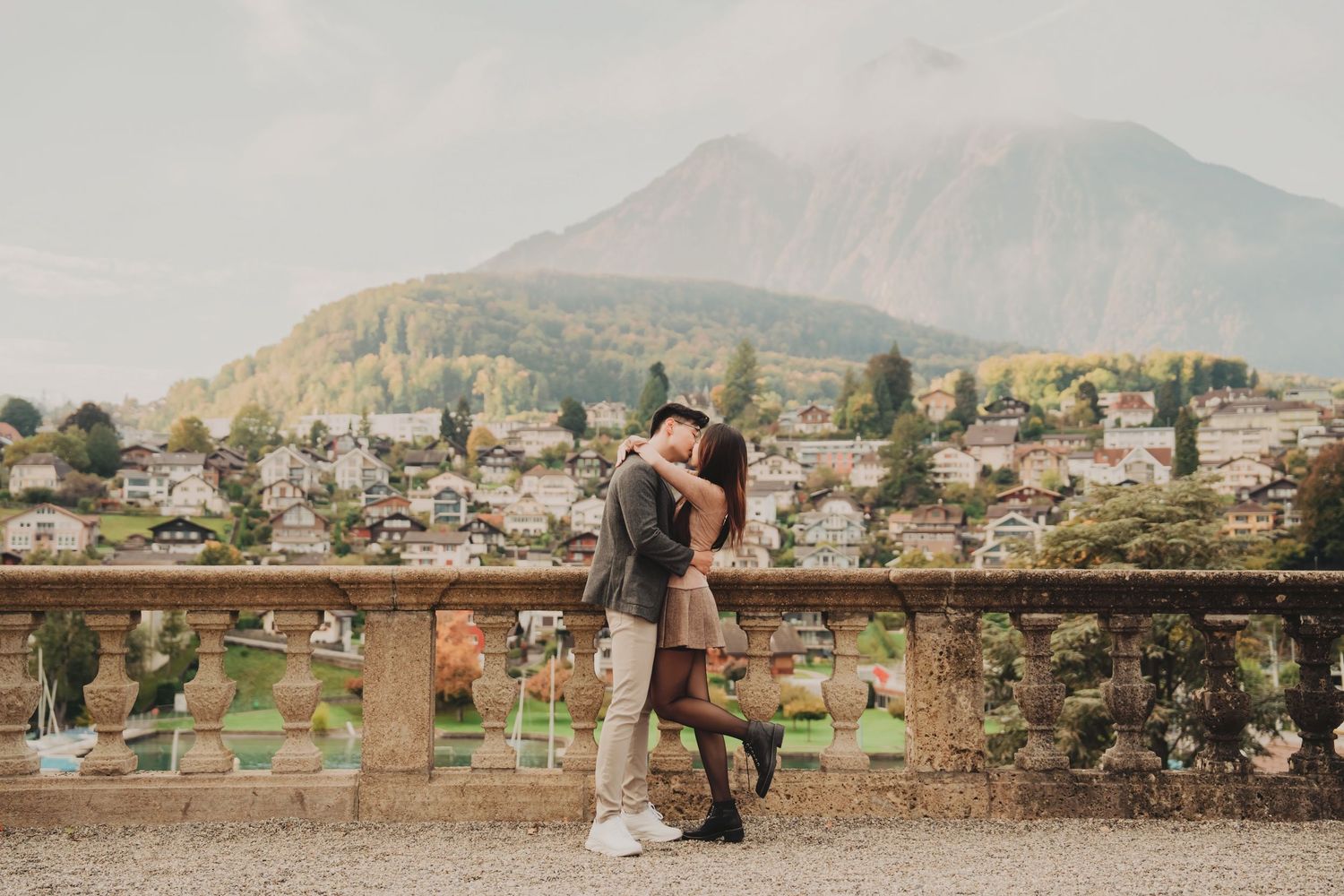 Couple sharing a romantic kiss on a scenic terrace with Alpine village and mountain backdrop.