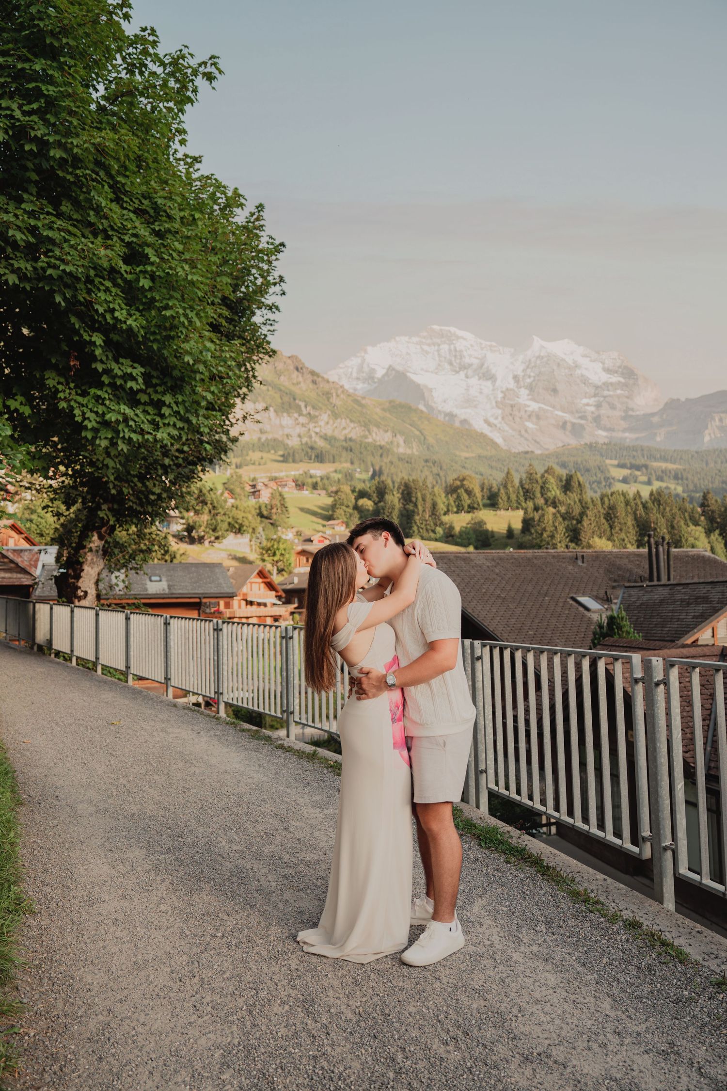 Couple sharing a tender kiss on a scenic alpine road surrounded by lush green trees and snow-capped Swiss mountains.