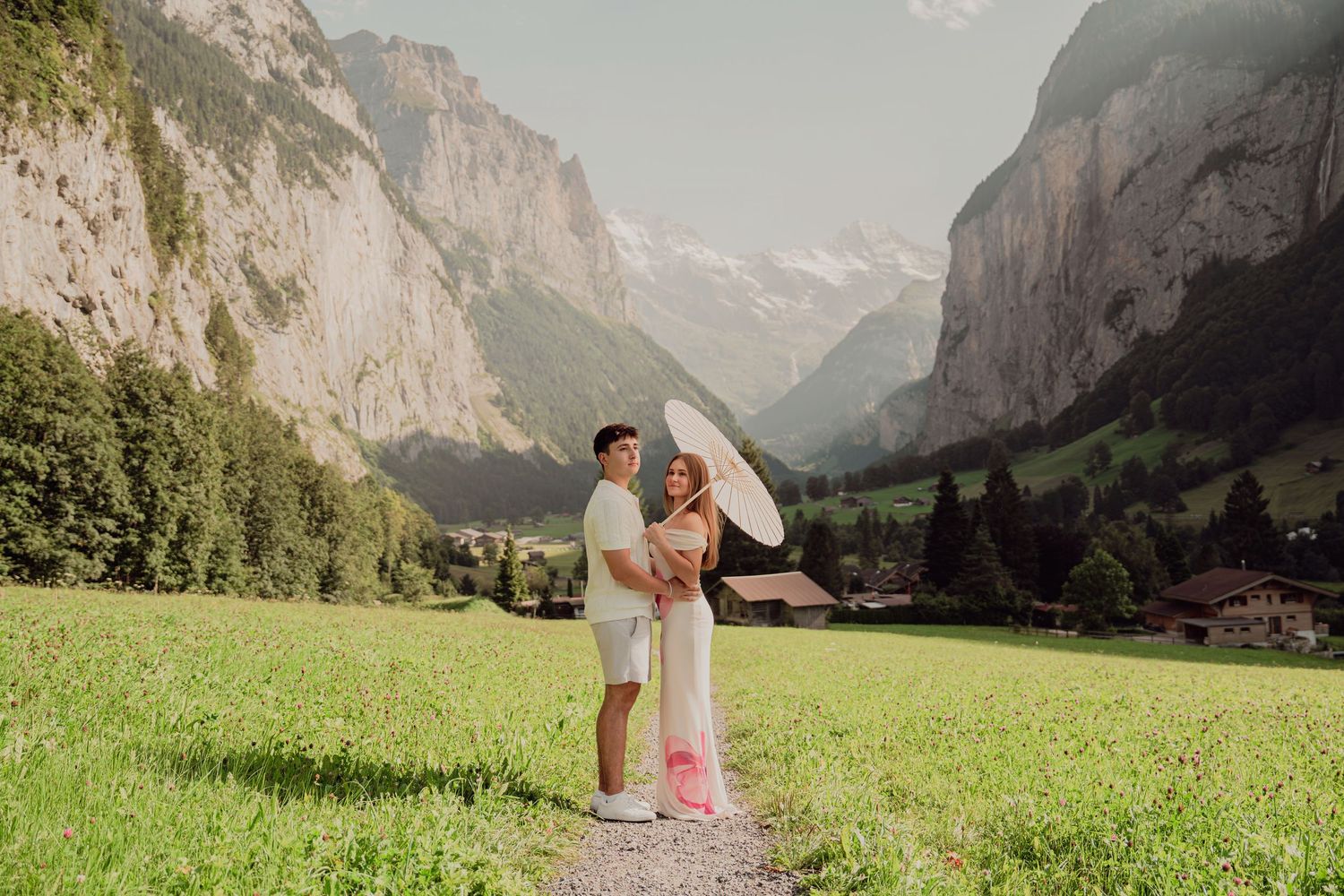 Couple standing close together under white parasol on a green meadow with majestic Swiss alpine valley backdrop.