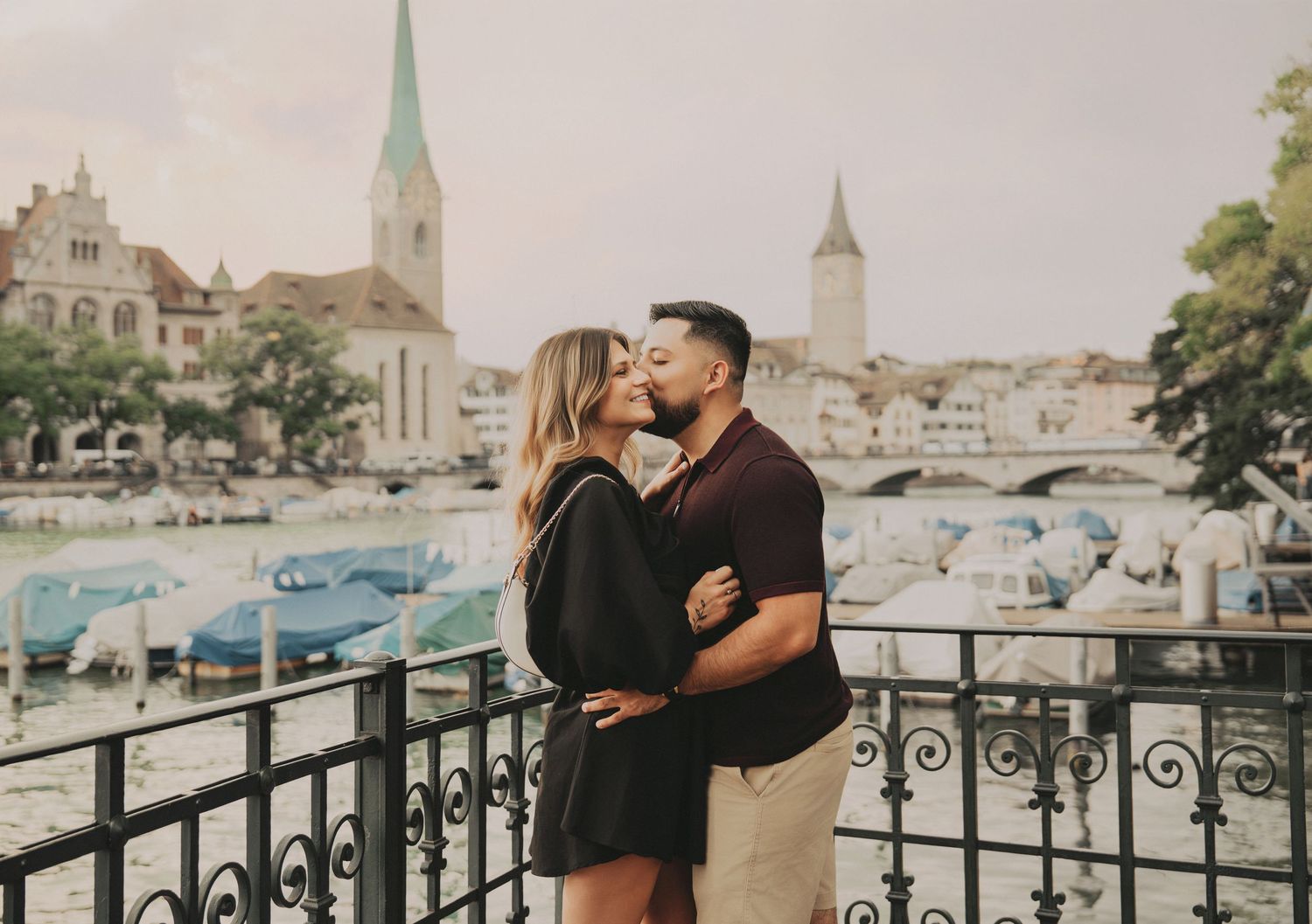 Couple kissing on a balcony with scenic Zurich river, boats, and historic church towers behind.