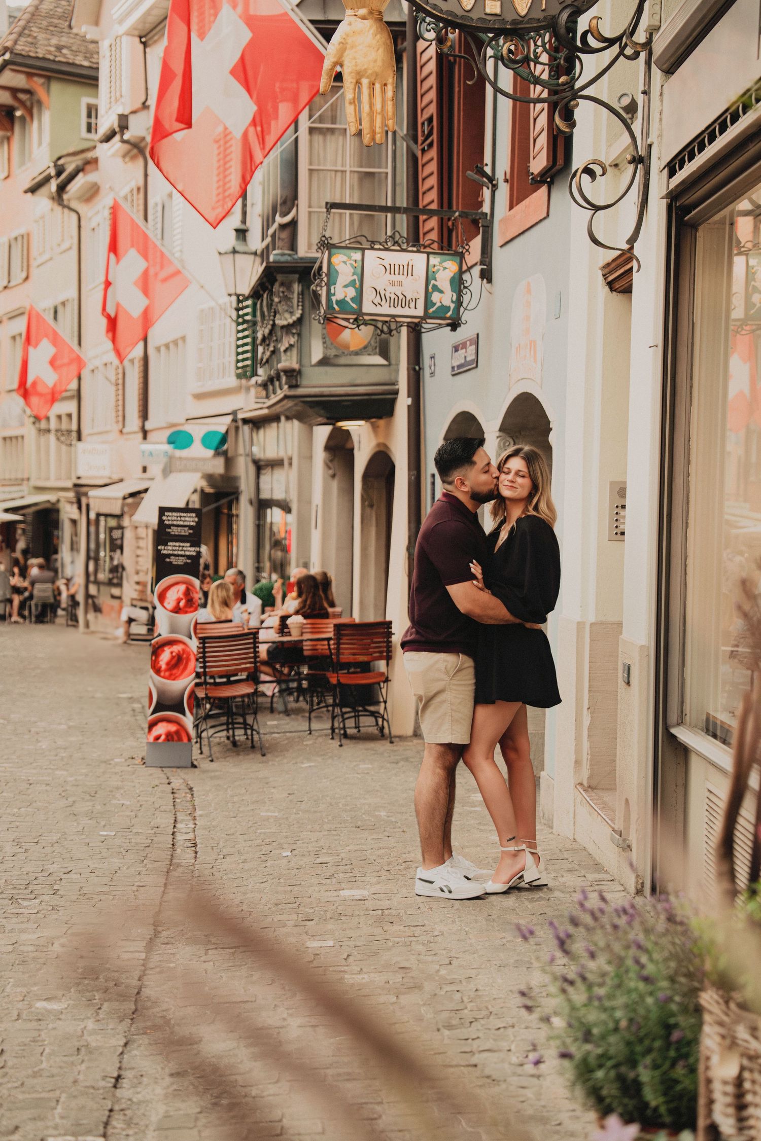 Couple embracing on charming Swiss street lined with red Swiss flags and cafes.