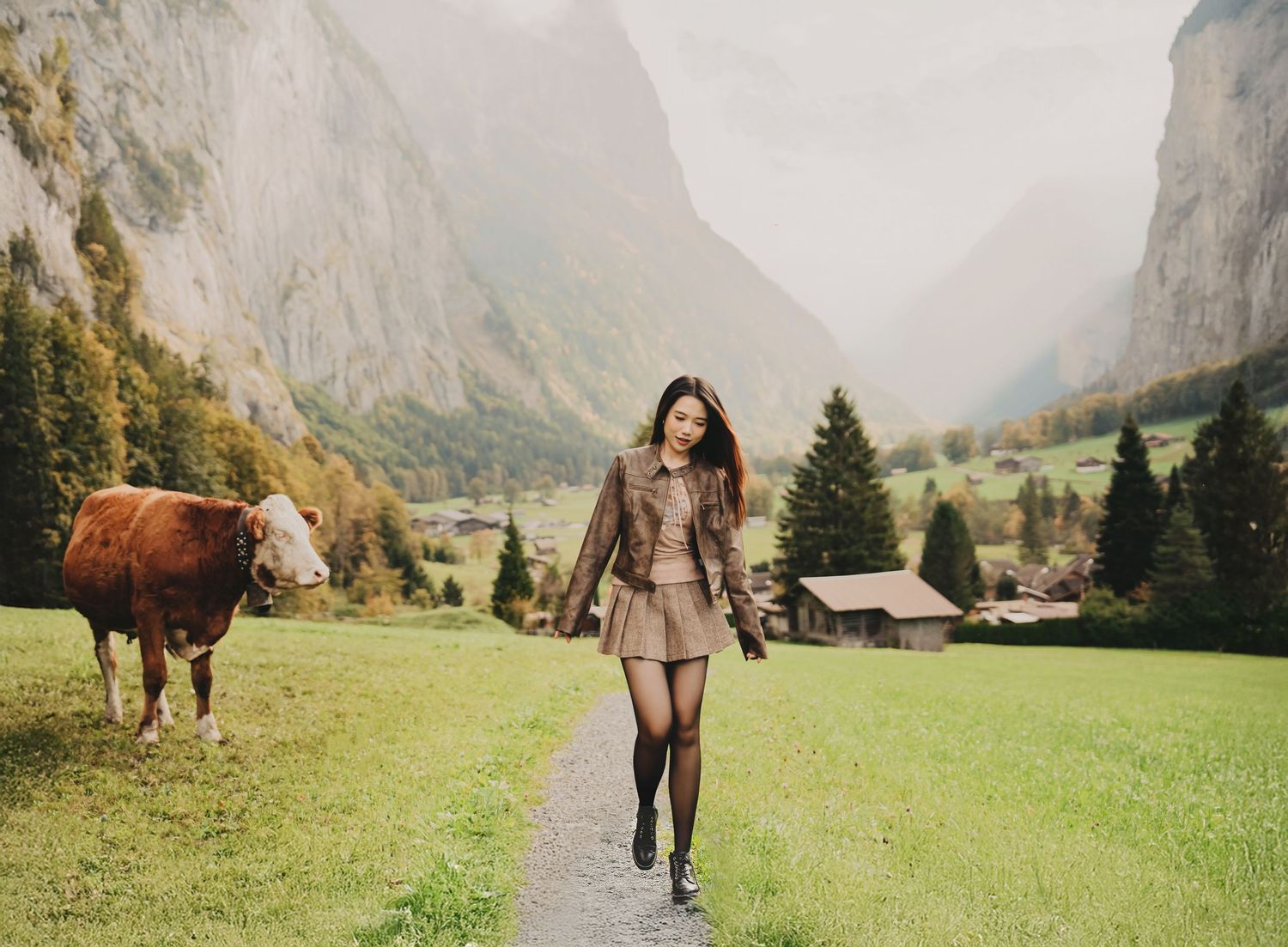 Woman in brown outfit walking on a path with a cow and Swiss alpine village in the background.
