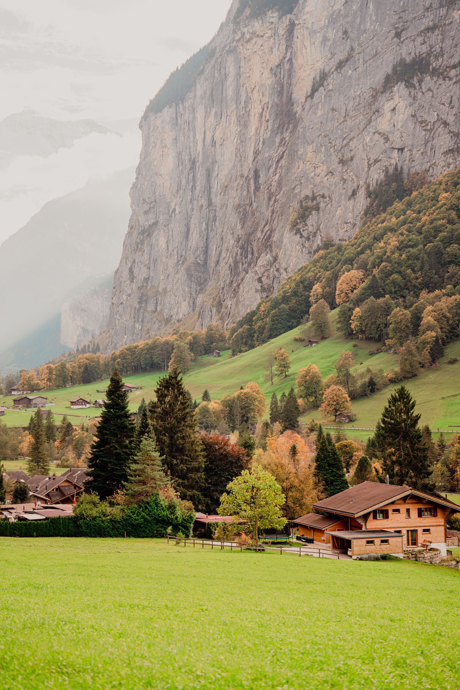 Charming Swiss alpine chalet surrounded by lush green meadows with dramatic rocky mountain cliffs in fog.