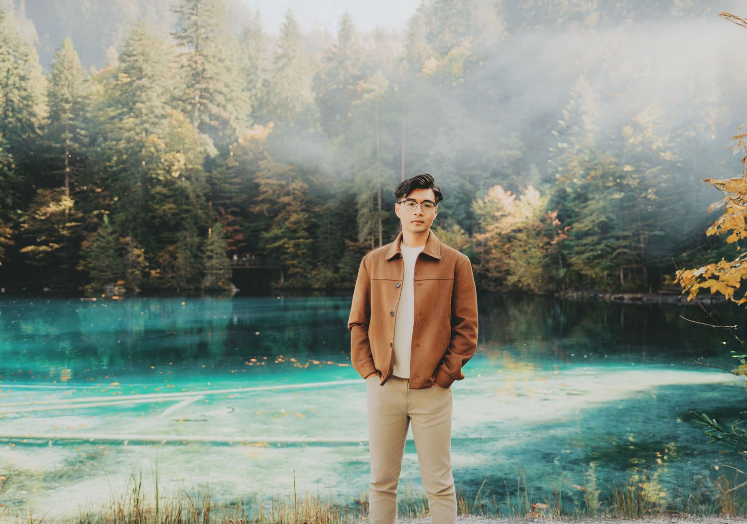 Young man in brown jacket stands before misty turquoise lake surrounded by tall pine trees in autumn.