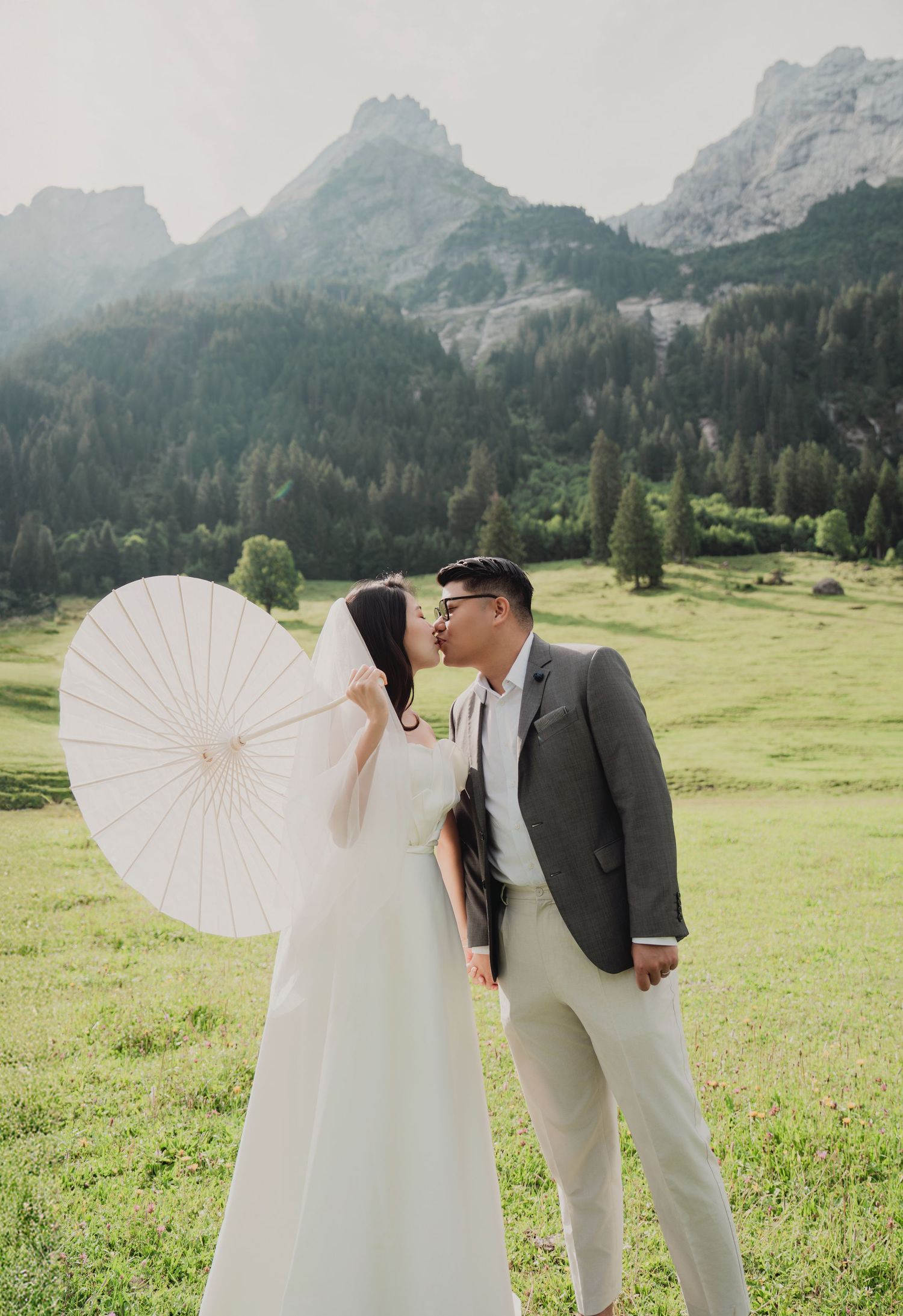 Bride and groom share romantic kiss in alpine setting, bride holding white parasol with dramatic mountain scenery.