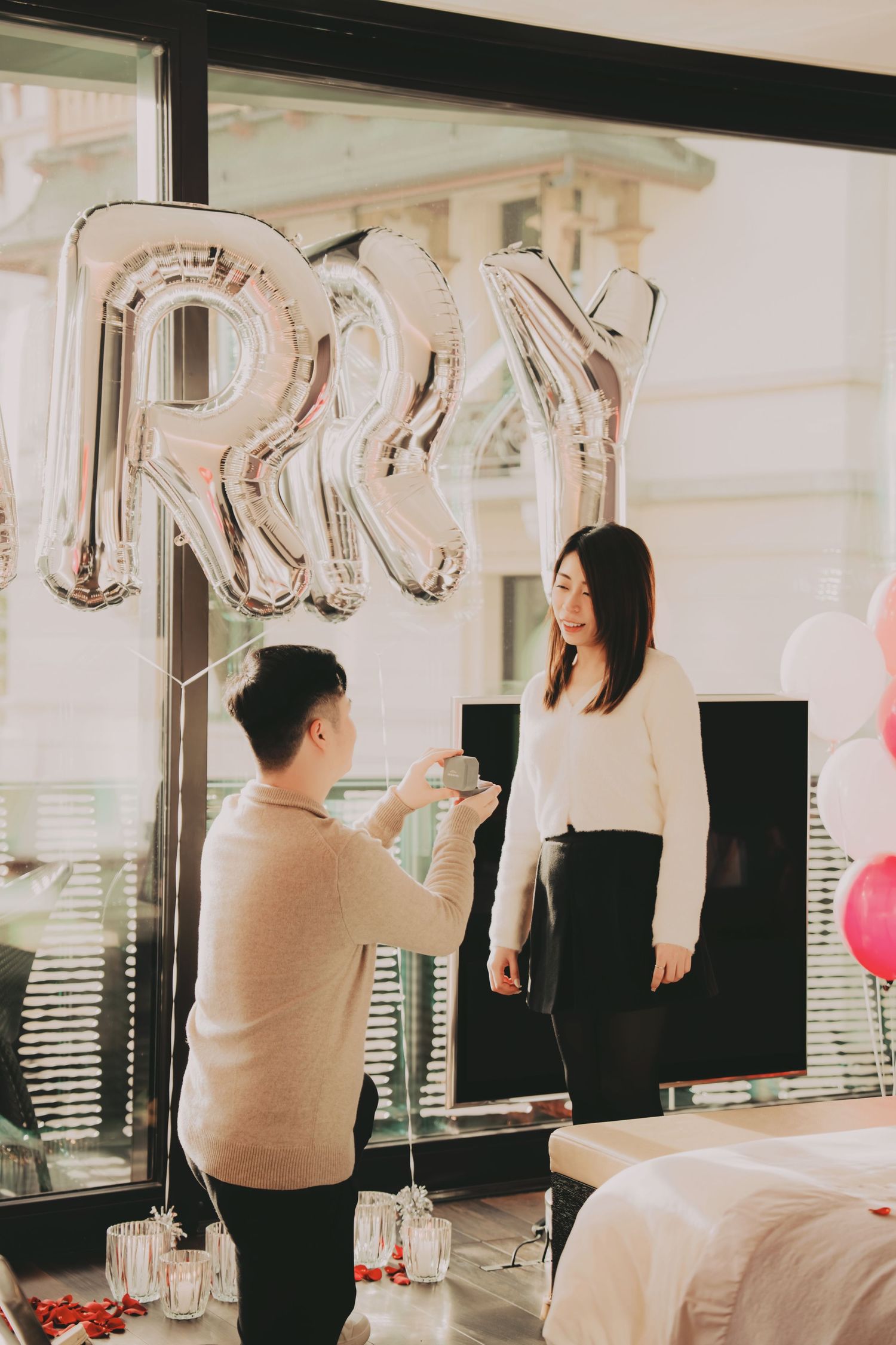 Romantic marriage proposal scene with man on one knee presenting ring box under silver letter balloons spelling MARRY.
