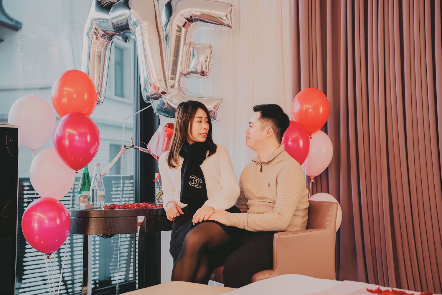 Couple sitting together surrounded by pink and red balloons in a romantic hotel room celebration.