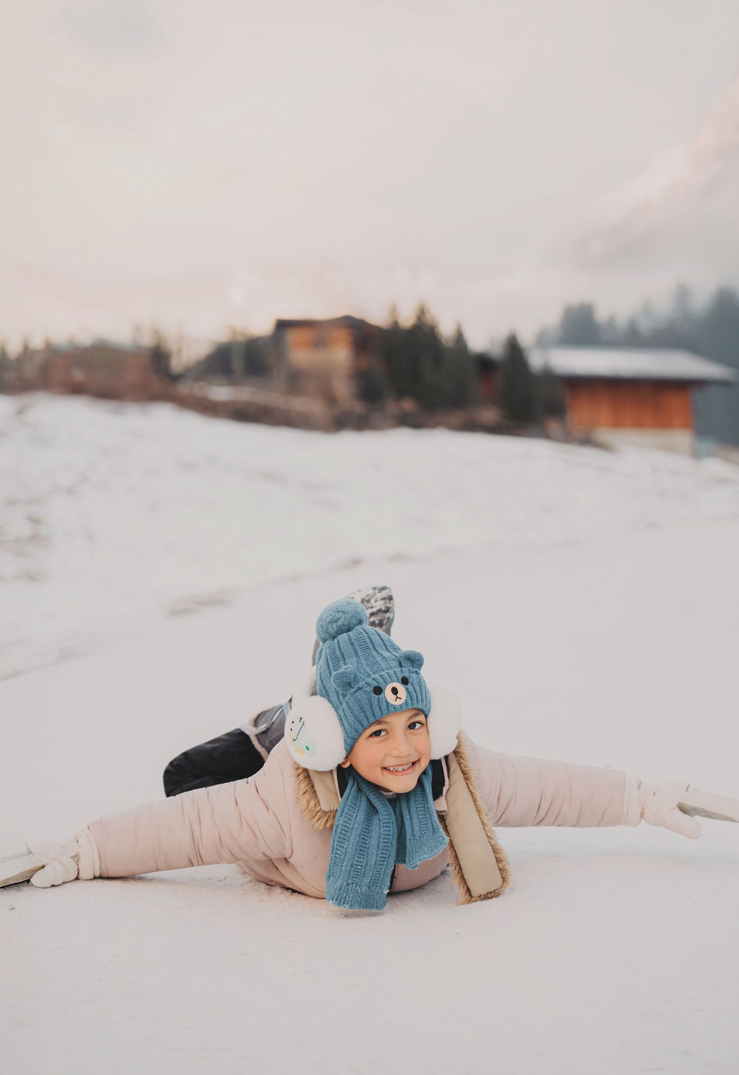 Happy child in blue bear hat and pink outfit doing splits on snowy ground smiling at camera.