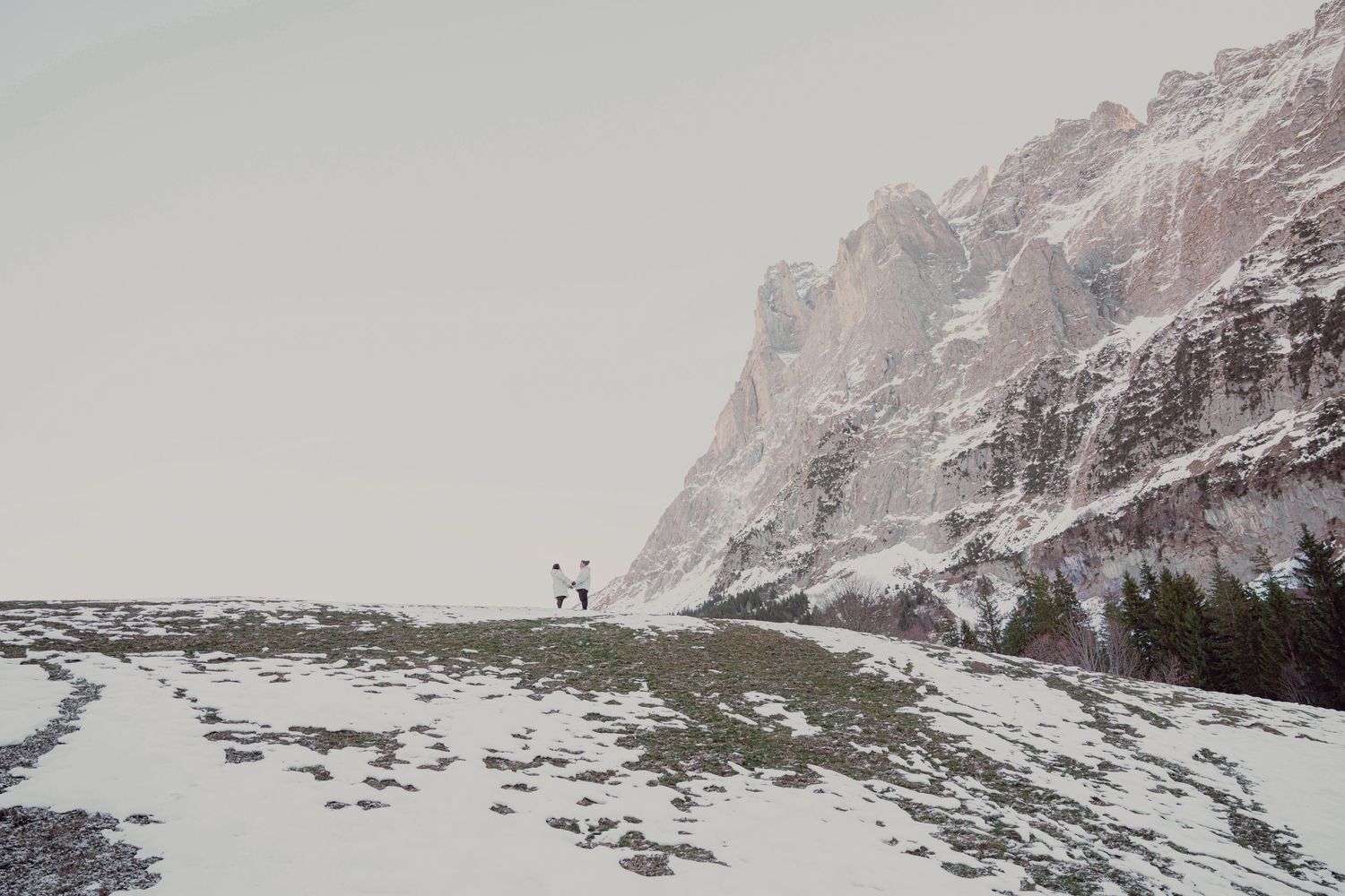 Two people standing on snowy hillside beside dramatic snow-covered rocky mountain cliff.