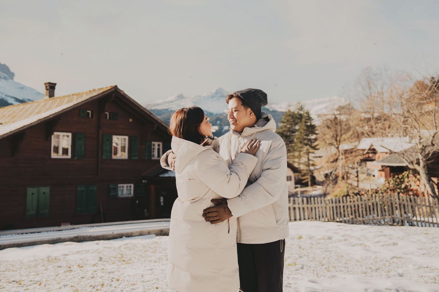 Couple hugging near rustic wooden chalet in snowy Swiss mountain village at sunset.