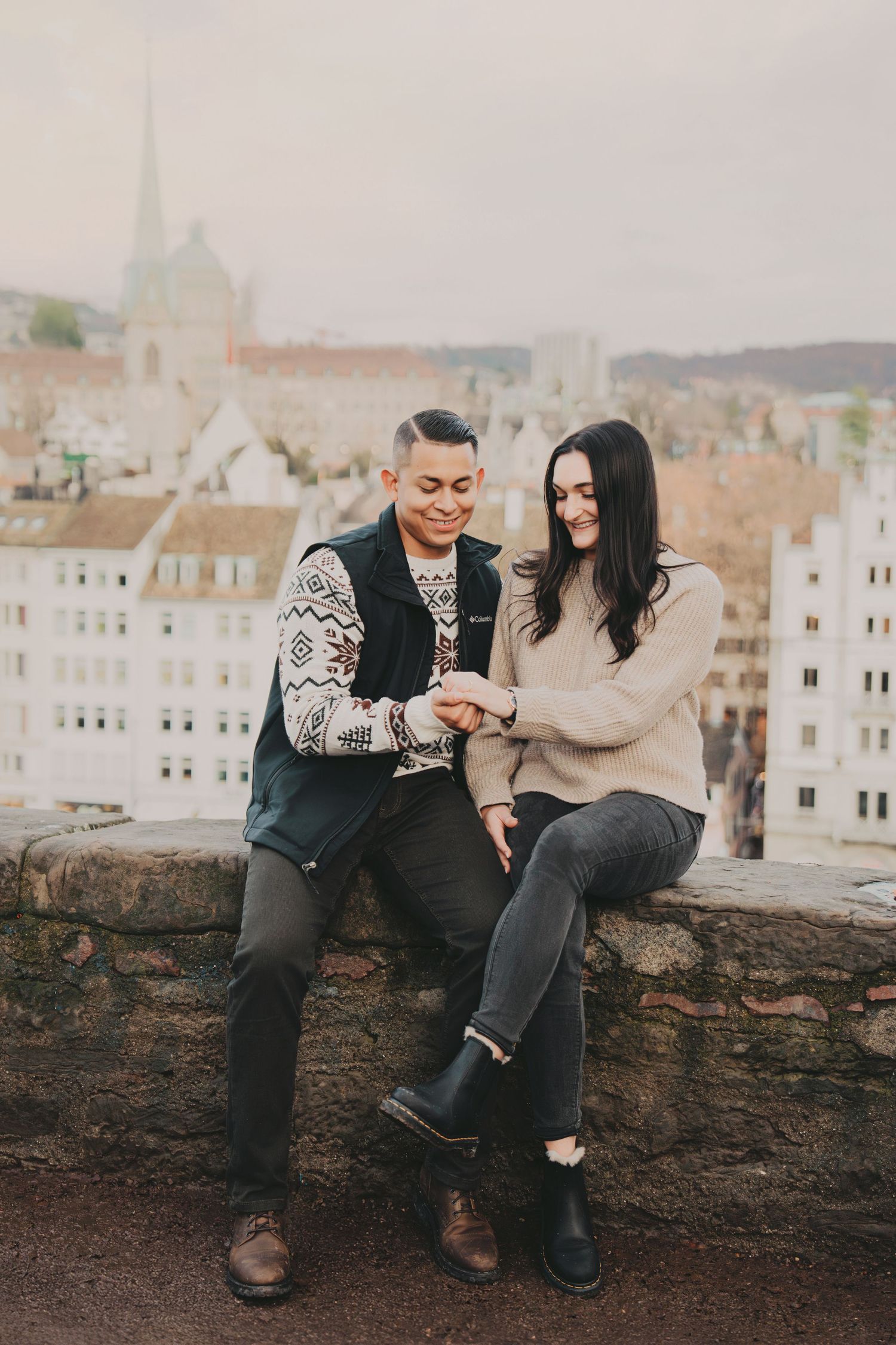 Smiling couple sitting on a stone wall admiring an engagement ring with city views.