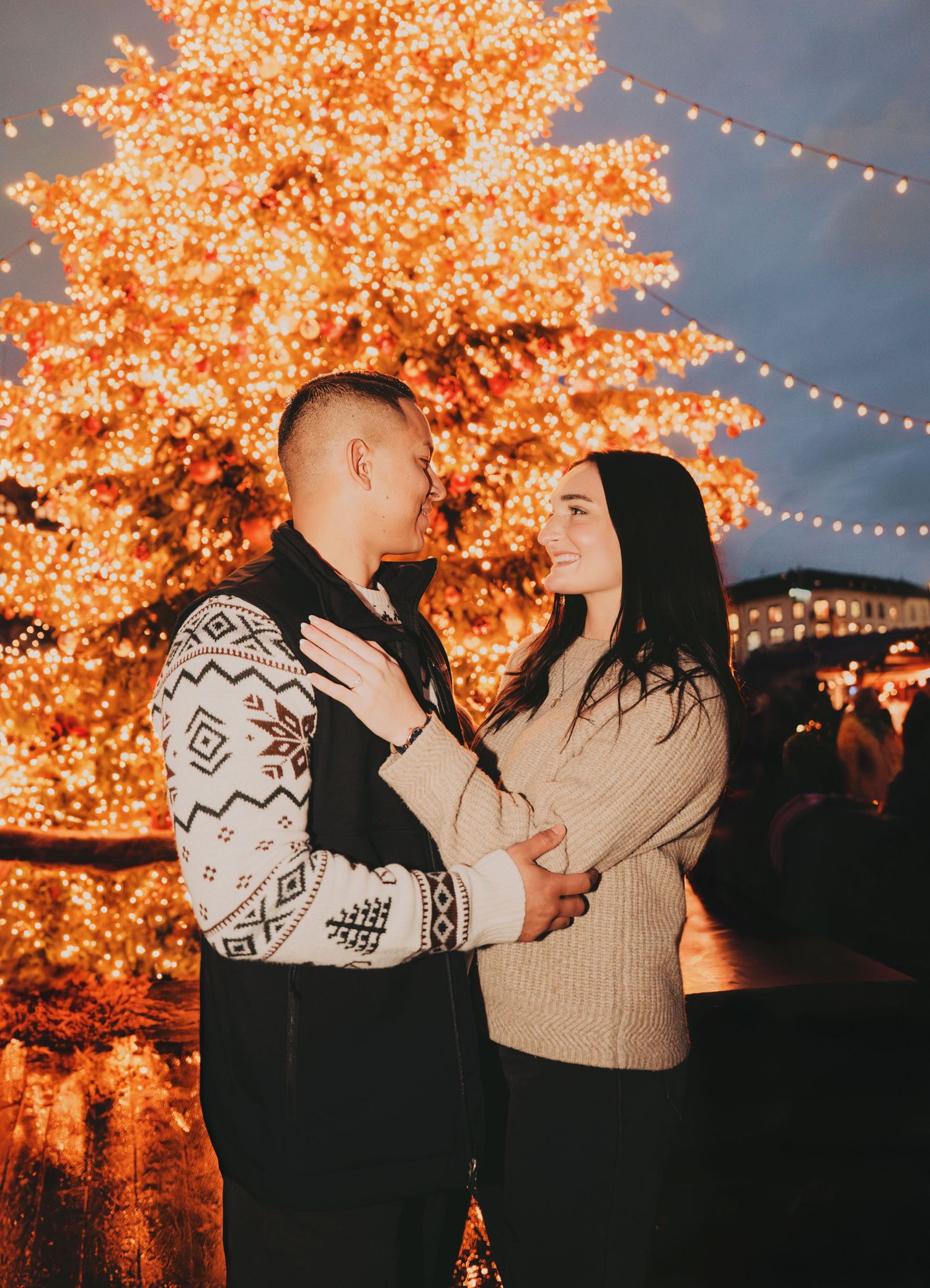 Couple embracing near a glowing Christmas tree with warm golden lights at a festive outdoor holiday market.
