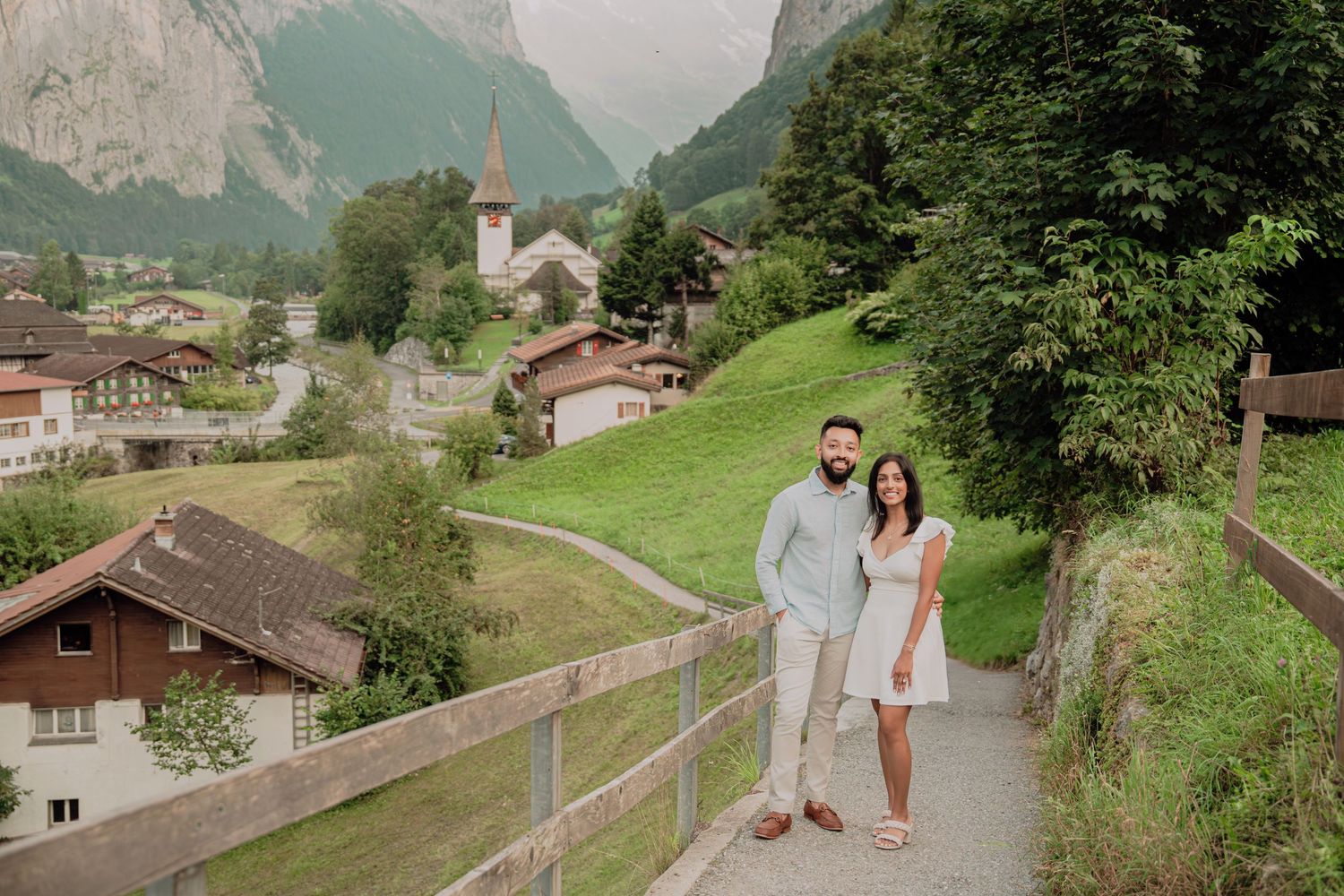 Smiling couple posing on walkway above charming Swiss village with church steeple and dramatic mountain cliffs in background.