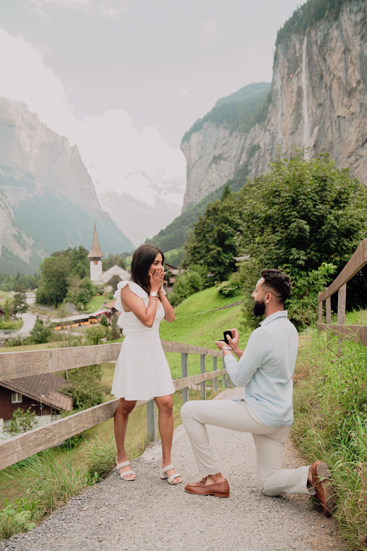 Man proposes on one knee to surprised woman covering her mouth in Lauterbrunnen, Switzerland.
