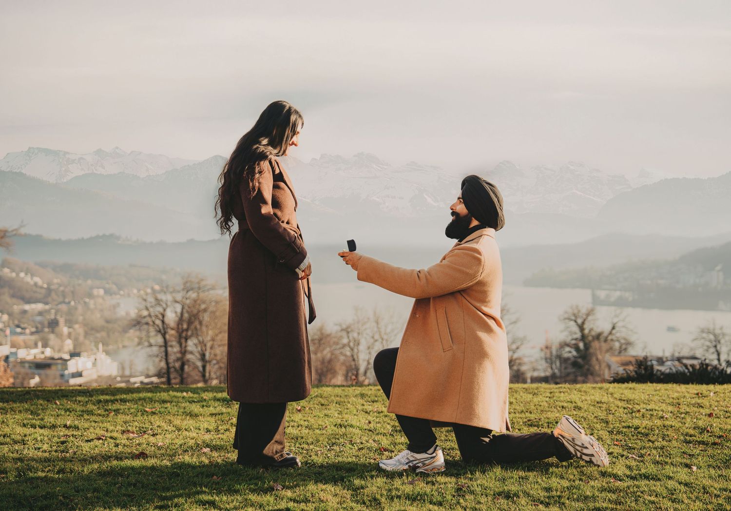 Man kneeling on one knee proposing to woman with ring box on a grassy hill with mountain views.
