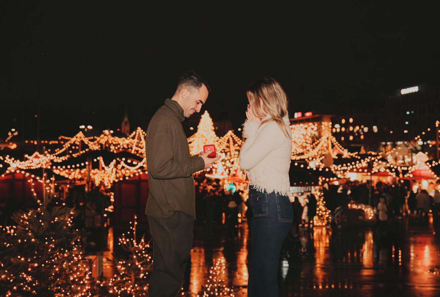 Man proposing to woman at romantic outdoor Christmas market with festive lights.