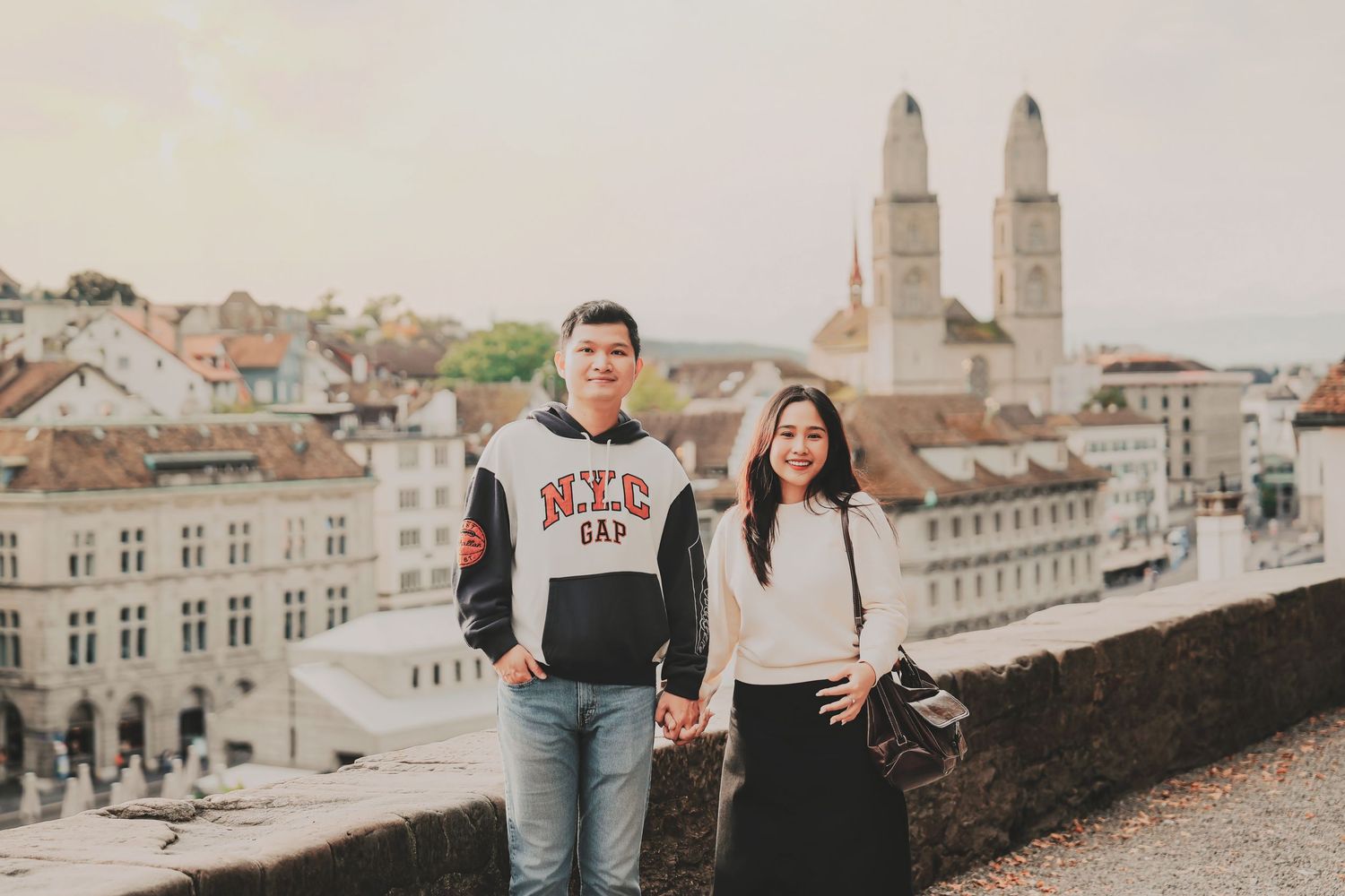 A couple holds hands and smiles with Zurich's iconic Grossmünster twin towers visible in the background.
