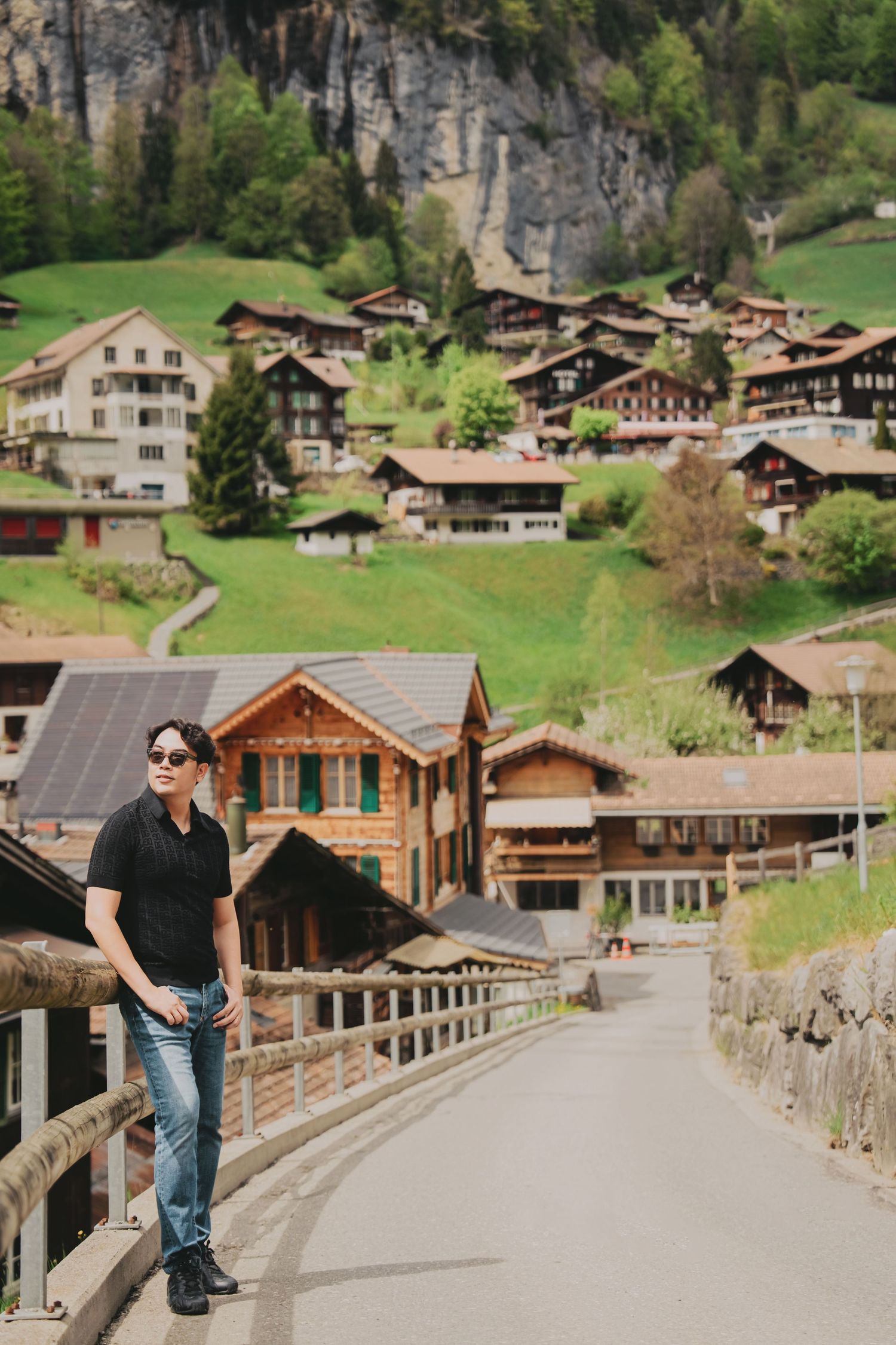 Man in black shirt and jeans posing on bridge with charming Swiss alpine village hillside behind.