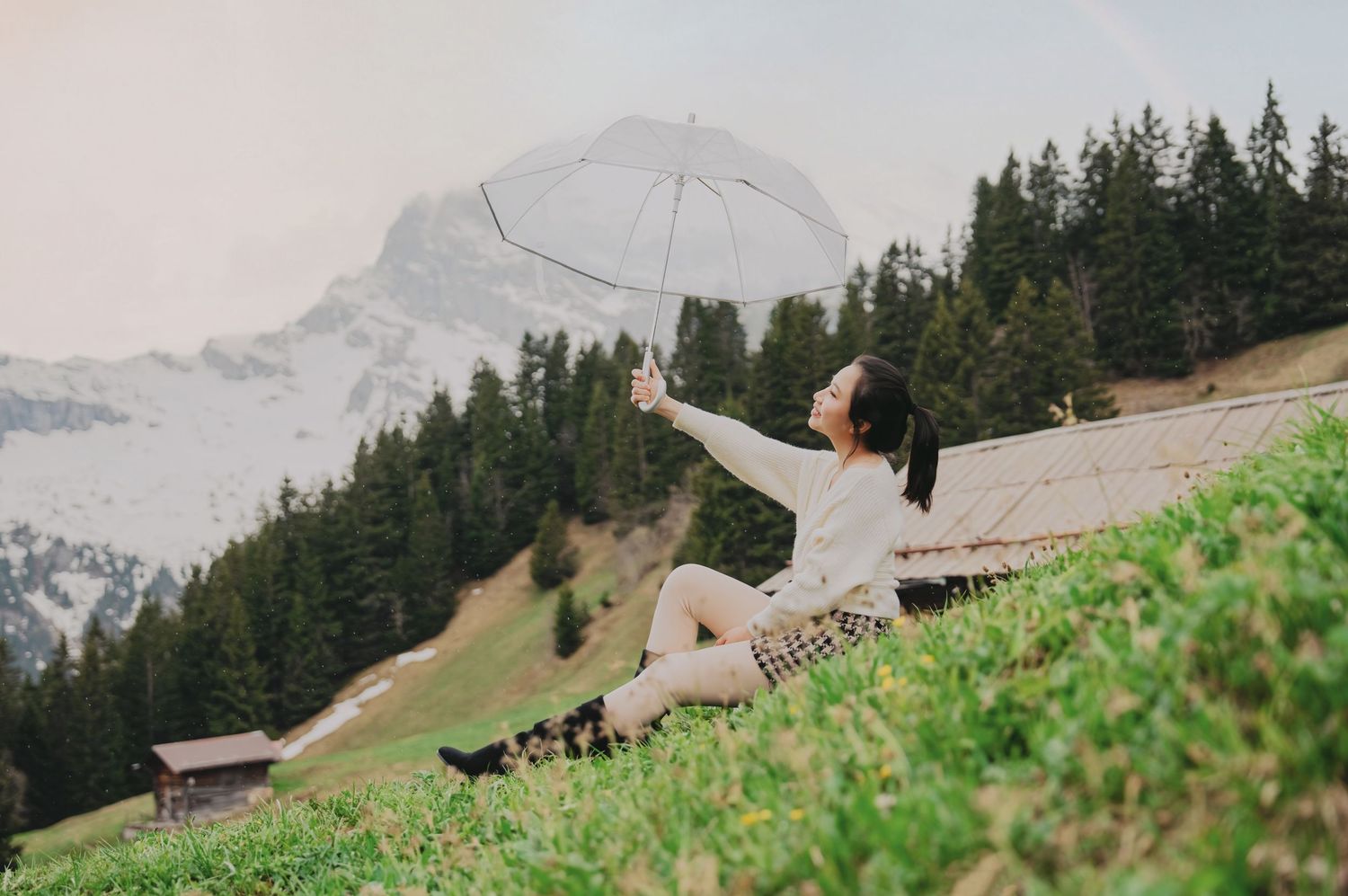 Young woman sitting on alpine hillside holding transparent umbrella with snowy mountain and pine trees behind.