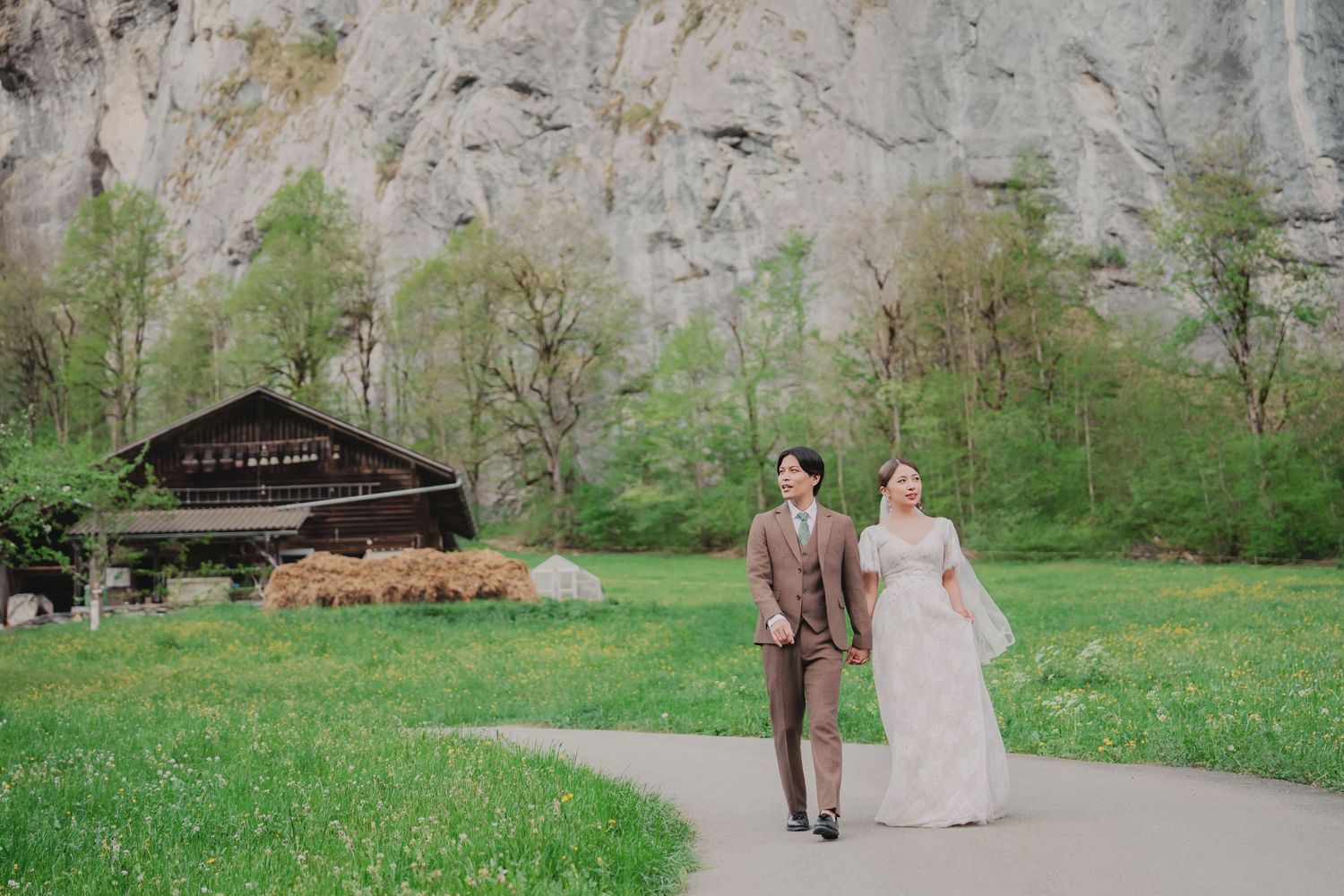 Bride and groom stroll along a path past a rustic alpine barn with dramatic rocky cliffs in Switzerland.
