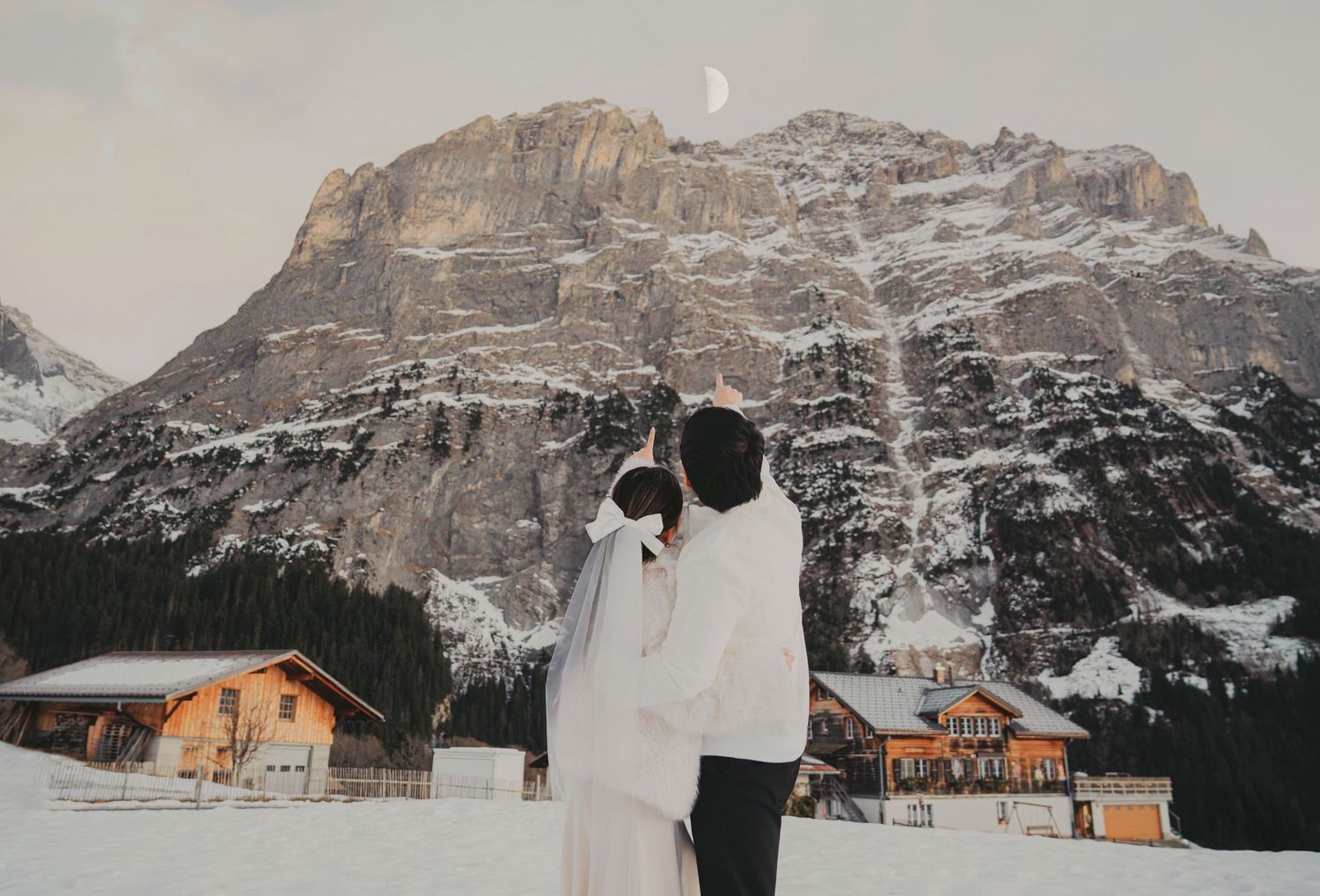 Couple in white attire facing snowy mountain landscape with moon visible at dusk in alpine village.